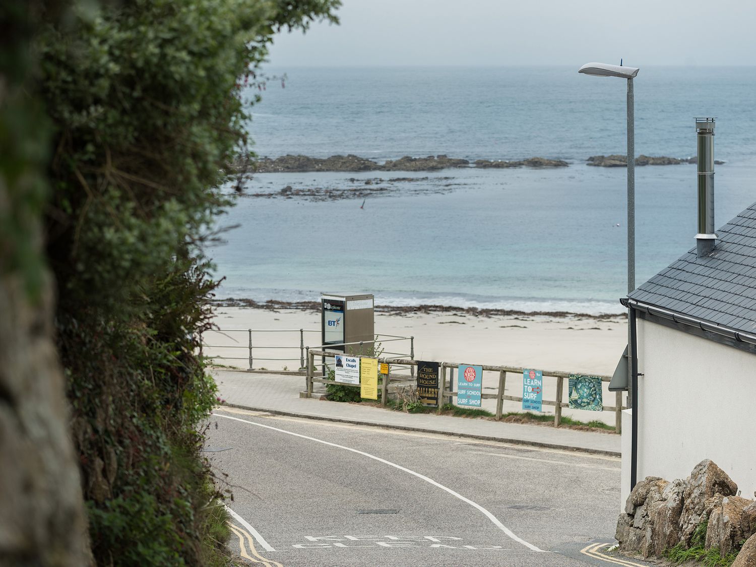 A coastal road with signs by a wooden fence and a sandy beach with ocean in the background at The Old Beach Store in Sennen Cove