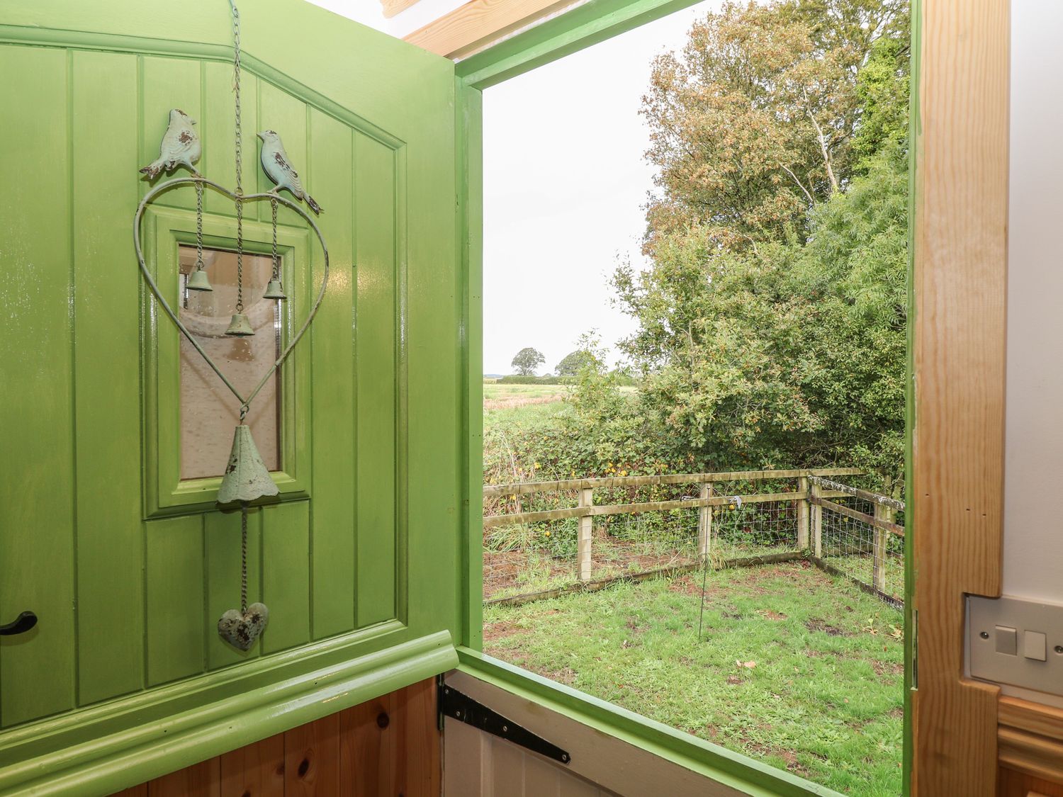 An open green door with a heart shaped hanging decoration overlooking a fenced garden with trees at The Old Mill Shepherd's Hut in Jacobstowe near Okehampton