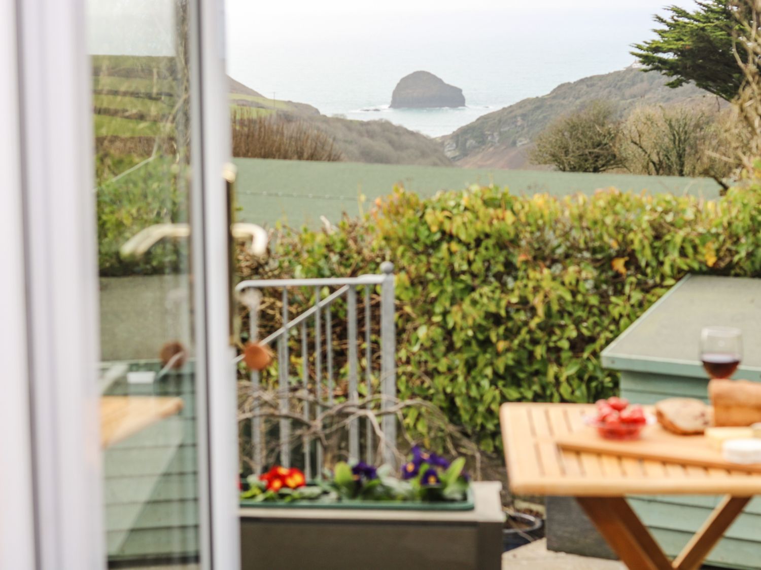 A view of a garden with a hedge and hills beyond with a rock in the sea seen through an open door with a wooden table holding bread and a glass of wine at Glenhaven in Trewarmett near Tintagel