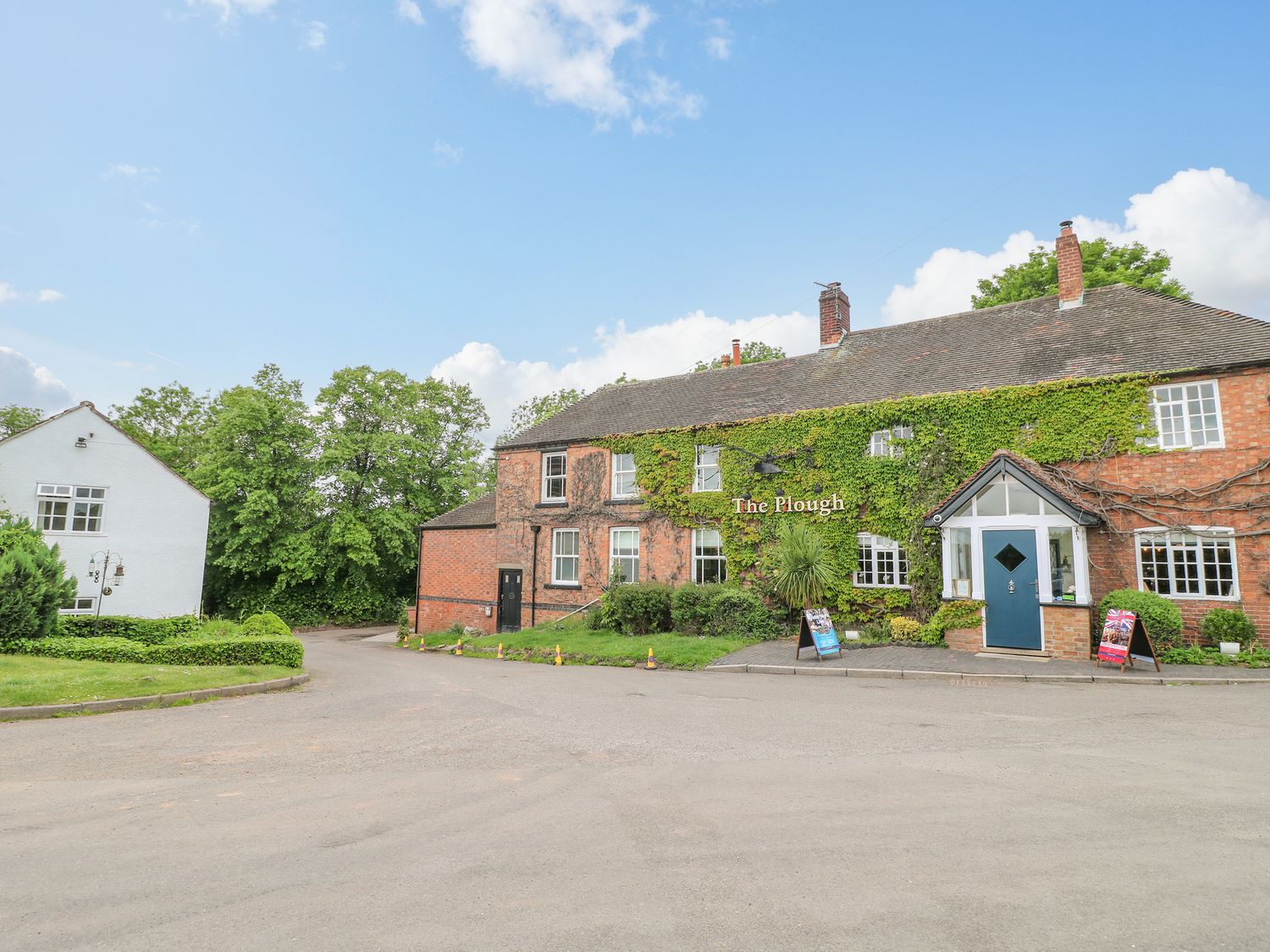 A brick building covered in ivy with a sign The Plough and a blue door near a white house and trees at Avenue Croft in Normanton-on-the-Wolds near Tollerton Nottinghamshire