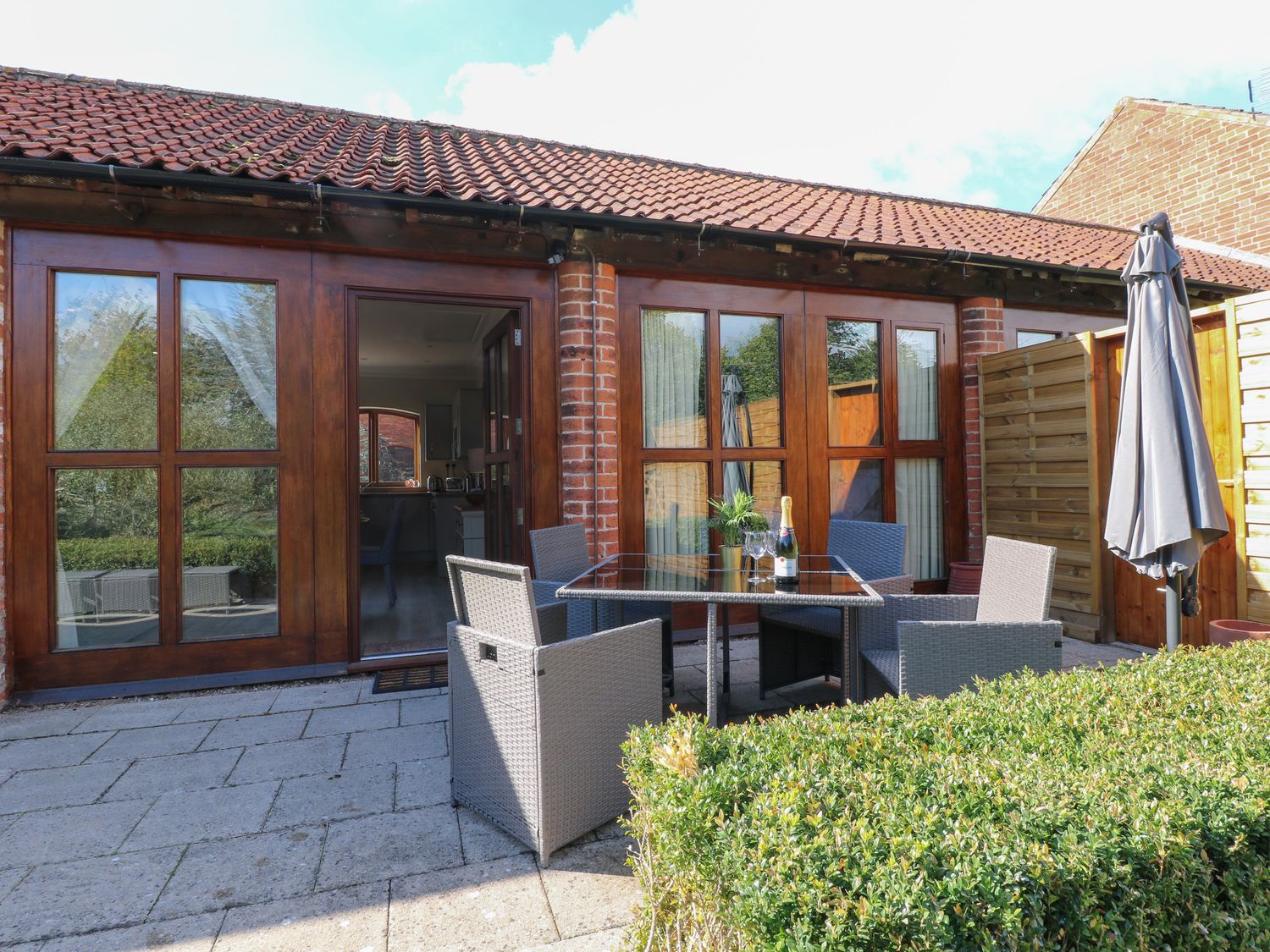 An outdoor patio area with a glass table, four chairs, a bottle of champagne, and an umbrella outside a building with wooden framed glass doors at Avenue Croft in Normanton-on-the-Wolds near Tollerton Nottinghamshire