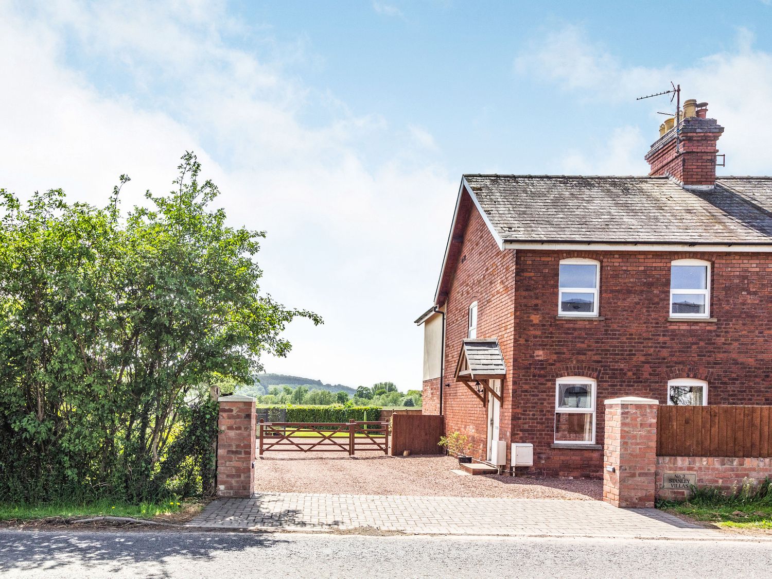 A brick house with a tiled roof and a gated driveway bordered by bushes at 2 Stanley Villas in Hereford