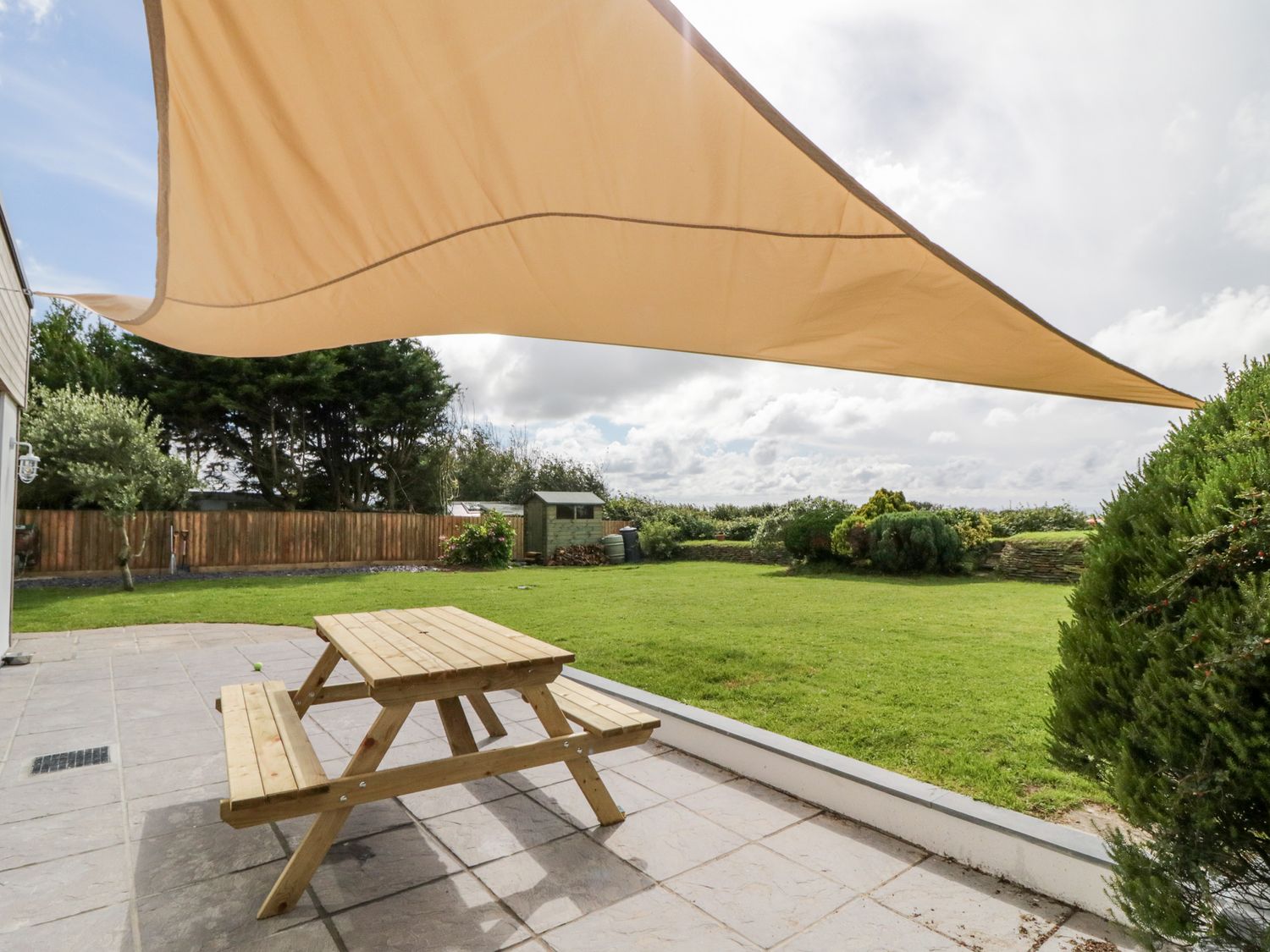A wooden picnic table on a tiled patio under a beige sunshade with a lawn and garden shed in the background at Fairways in Stibb near Bude