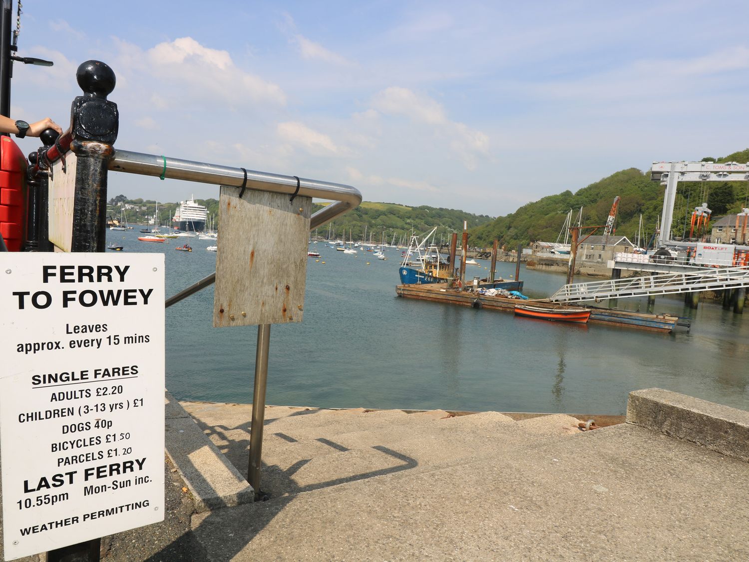 A ferry dock with a sign showing ferry fares beside a body of water with boats and green hills at The Old Chapel in Lanteglos Highway near Fowey