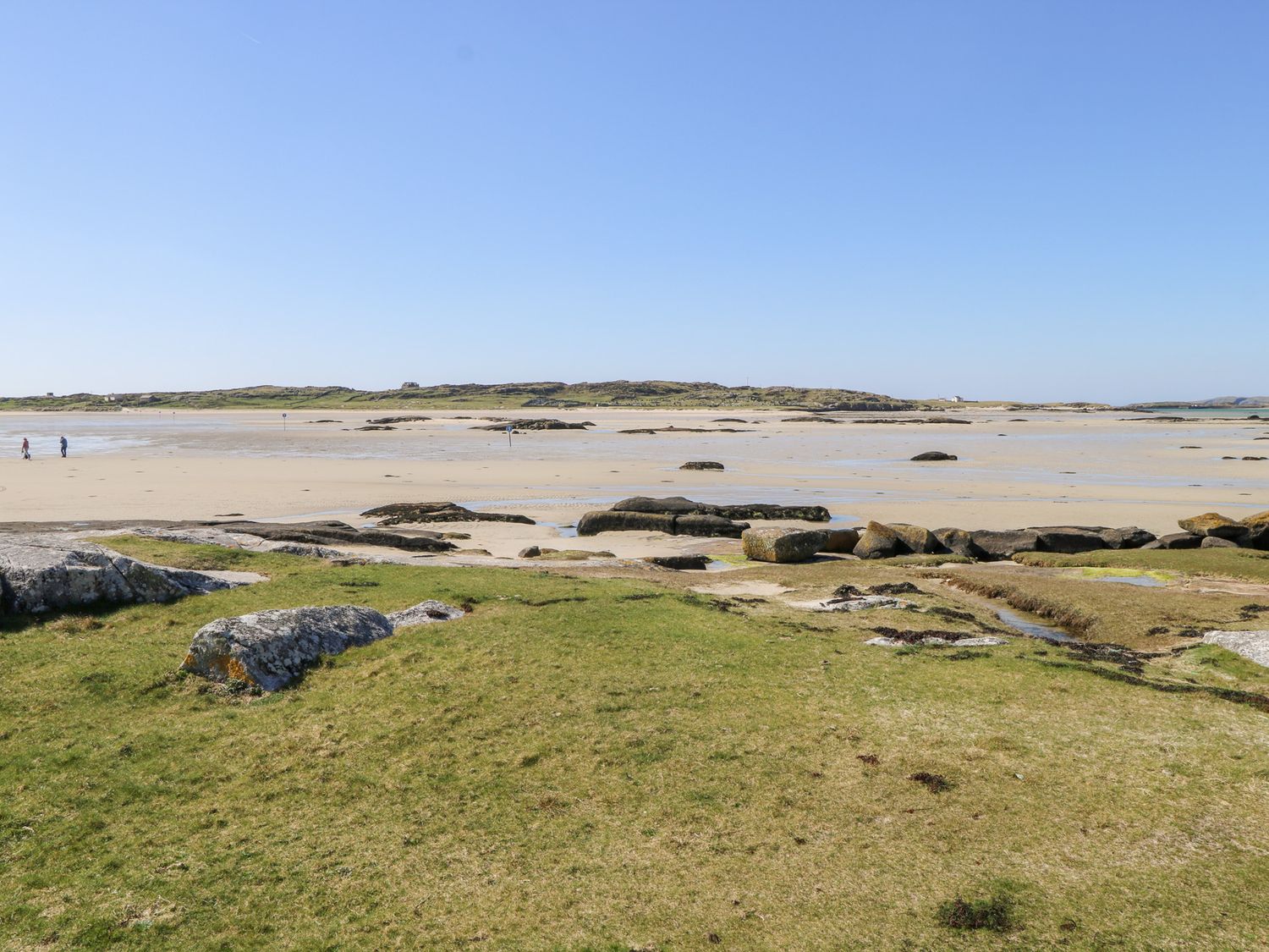 A sandy beach with rocks and grassy foreground and two people walking at Realt na Maidne in Claddaghduff County Galway