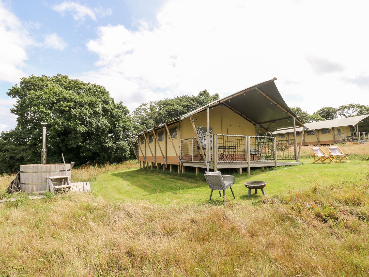 A raised tent structure with a covered porch and outdoor furniture on a grassy area near wooden hot tub and fire pit at Bumble @ Stars & Embers Glamping in Grampound
