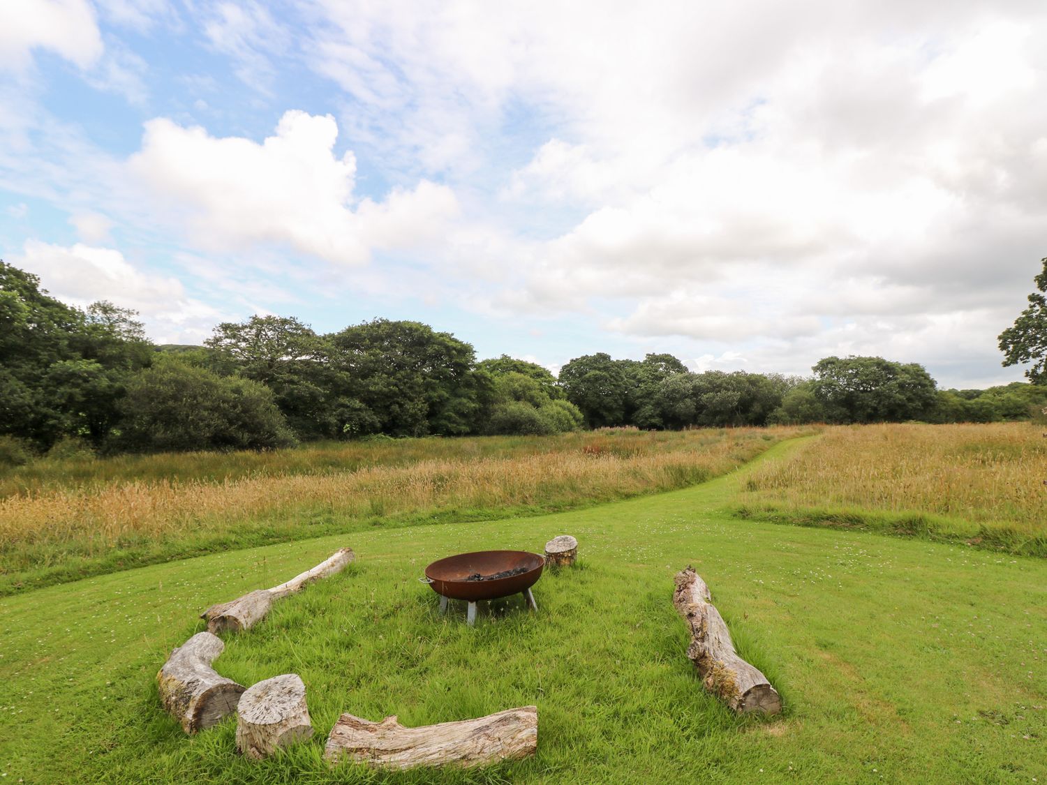 A grassy field with a fire pit surrounded by logs with trees and a cloudy sky in the background at Bumble @ Stars & Embers Glamping in Grampound