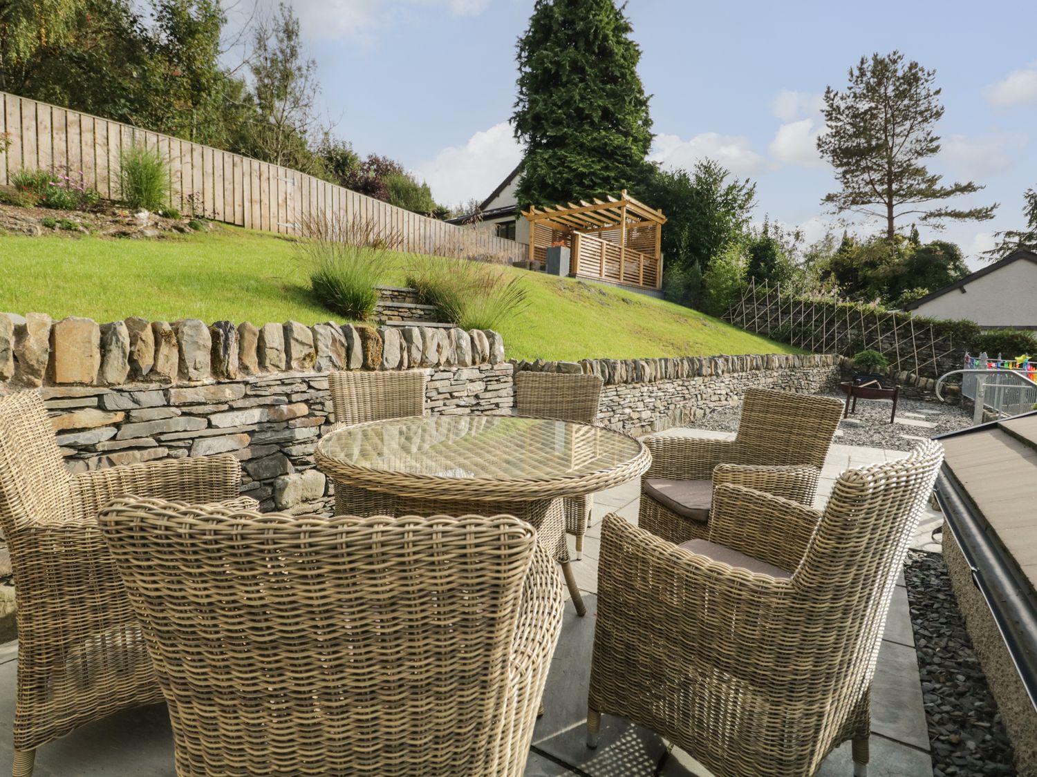 An outdoor seating area with six wicker chairs around a glass table on a patio next to a stone wall and grassy slope at Bishop Fell in Newby Bridge