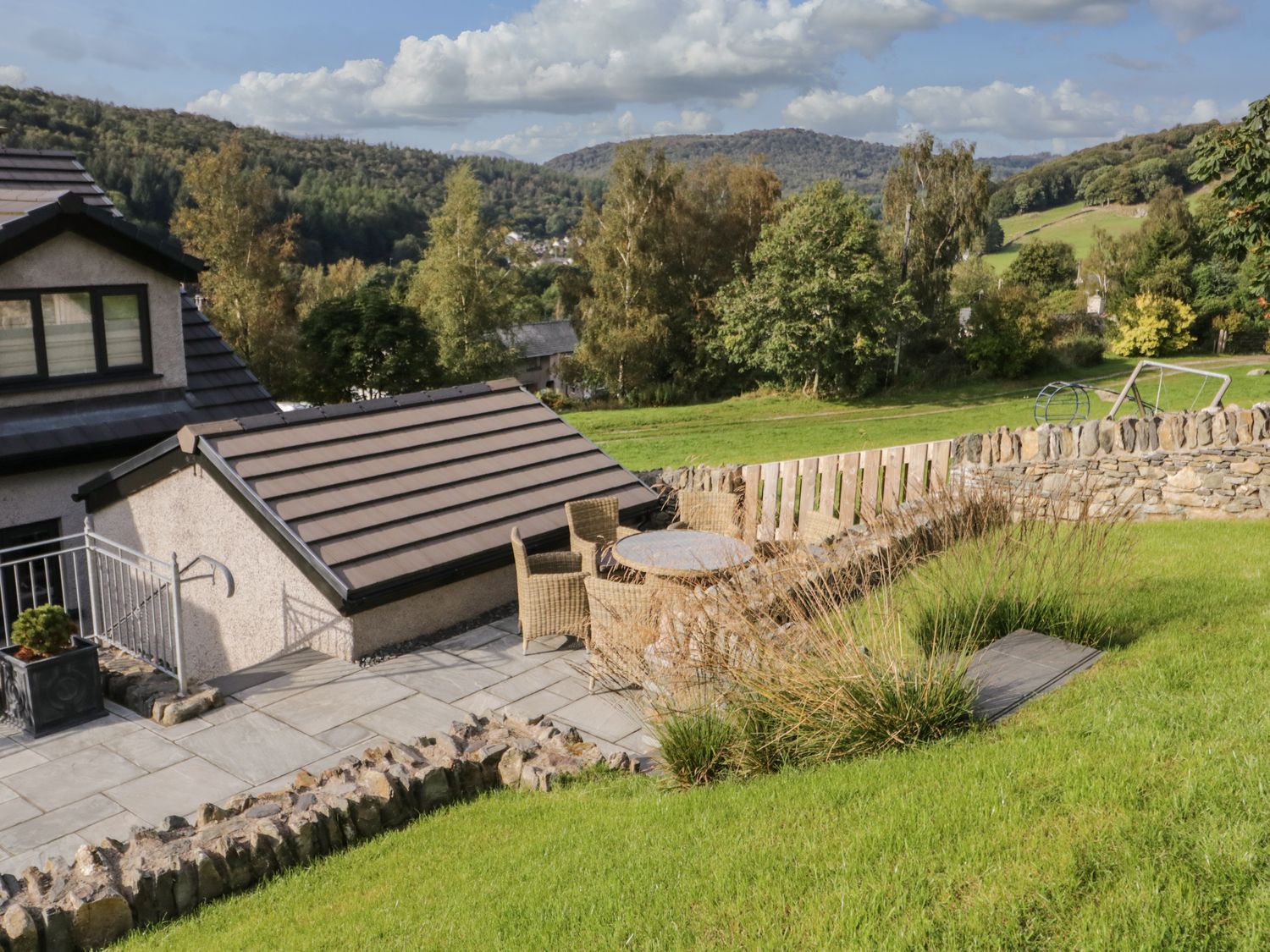 An outdoor patio area with a round table and wicker chairs next to a house with a tiled roof and green hills in the background at Bishop Fell in Newby Bridge