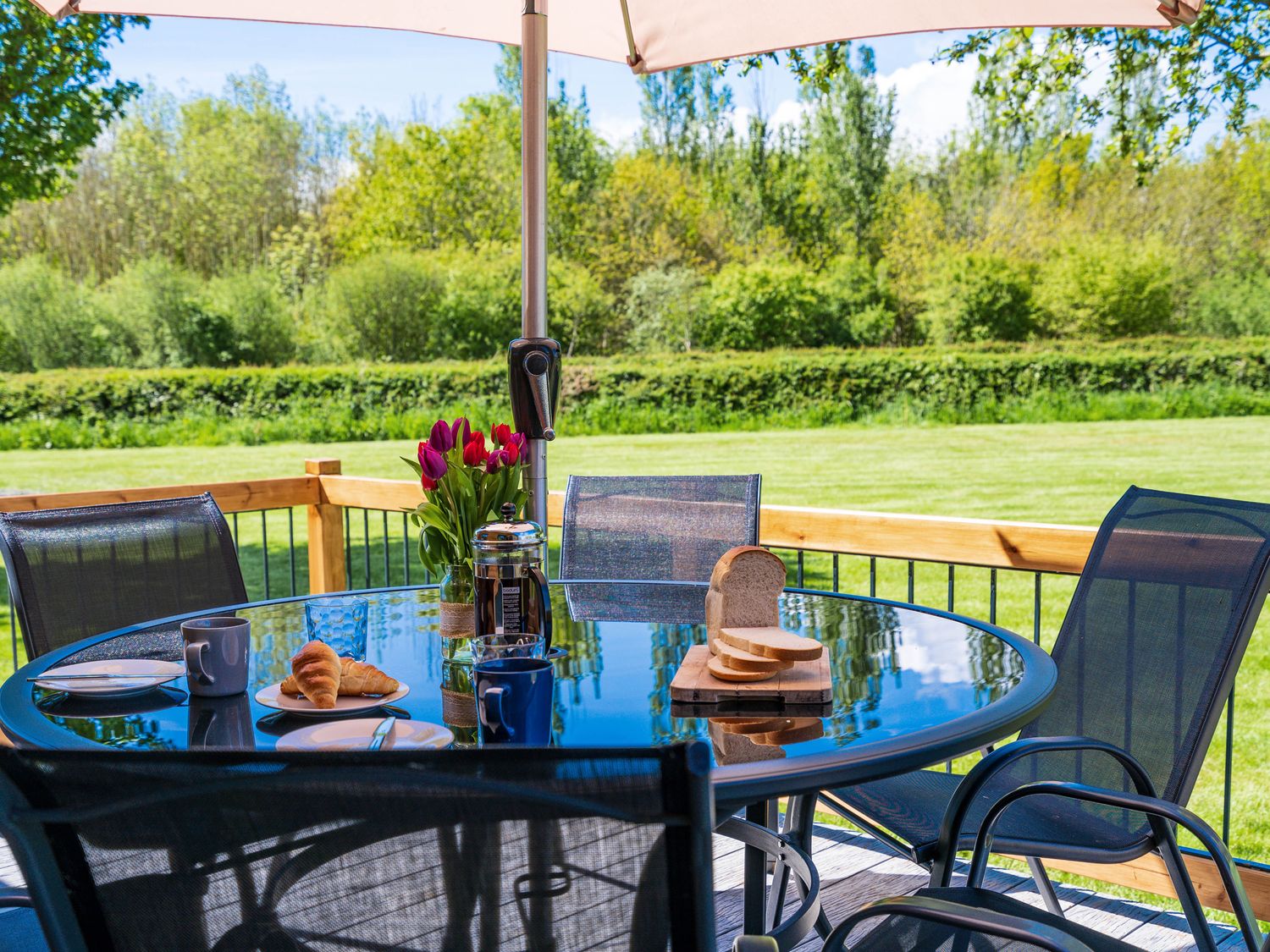 An outdoor glass table with croissants coffee mugs sliced bread and flowers under an umbrella at Top Style Orchard in Hallow