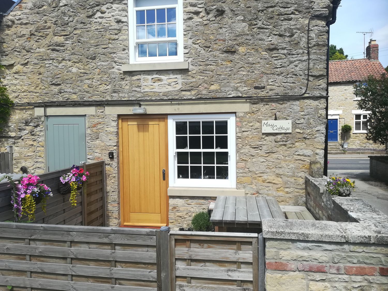 Stone cottage facade with wooden door window flower baskets and wooden fence at May Cottage in Snainton