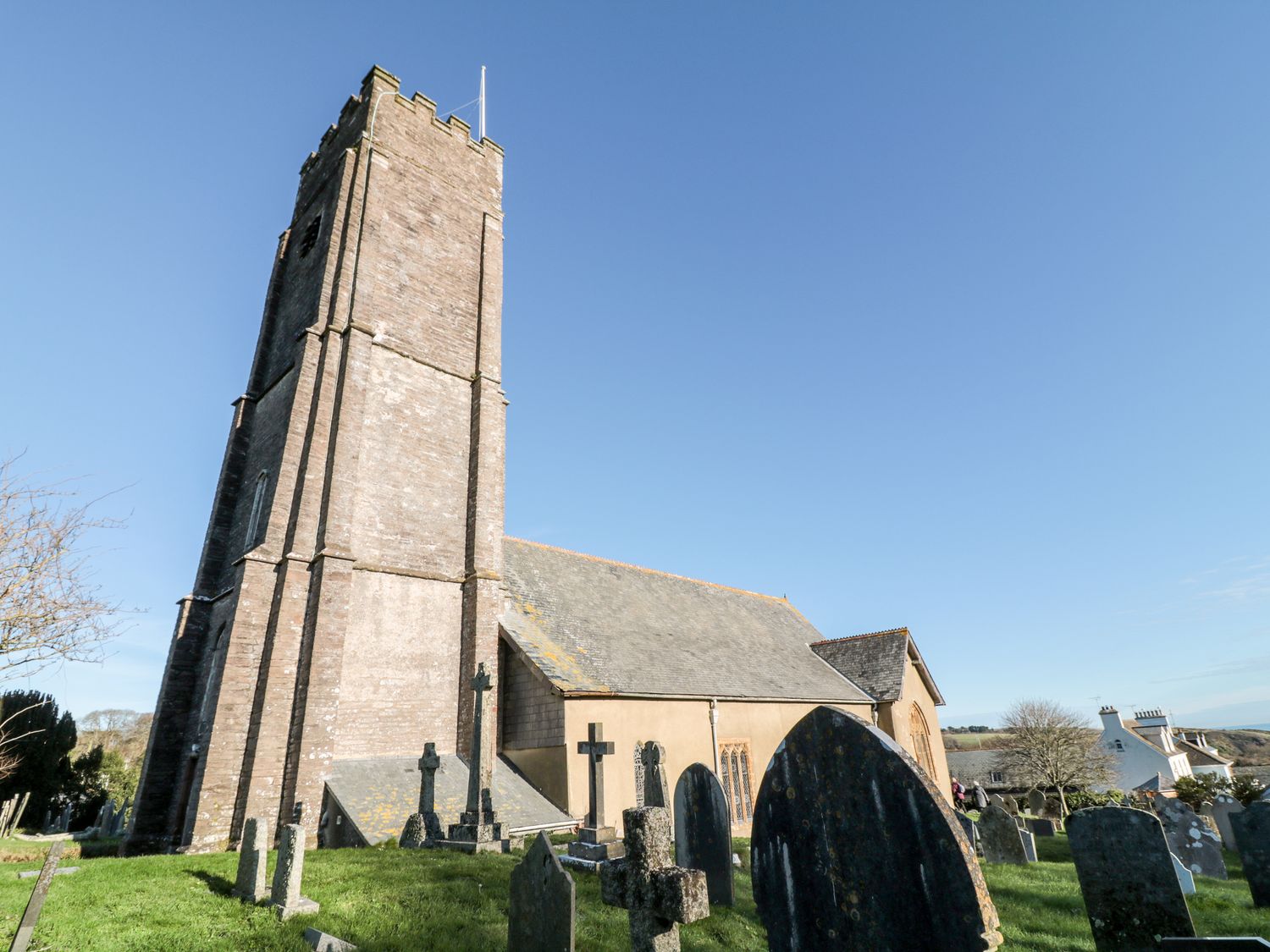 An old church building with a tall tower and gravestones in a graveyard with houses and fields in the background at Yew Tree Cottage in Stoke Fleming