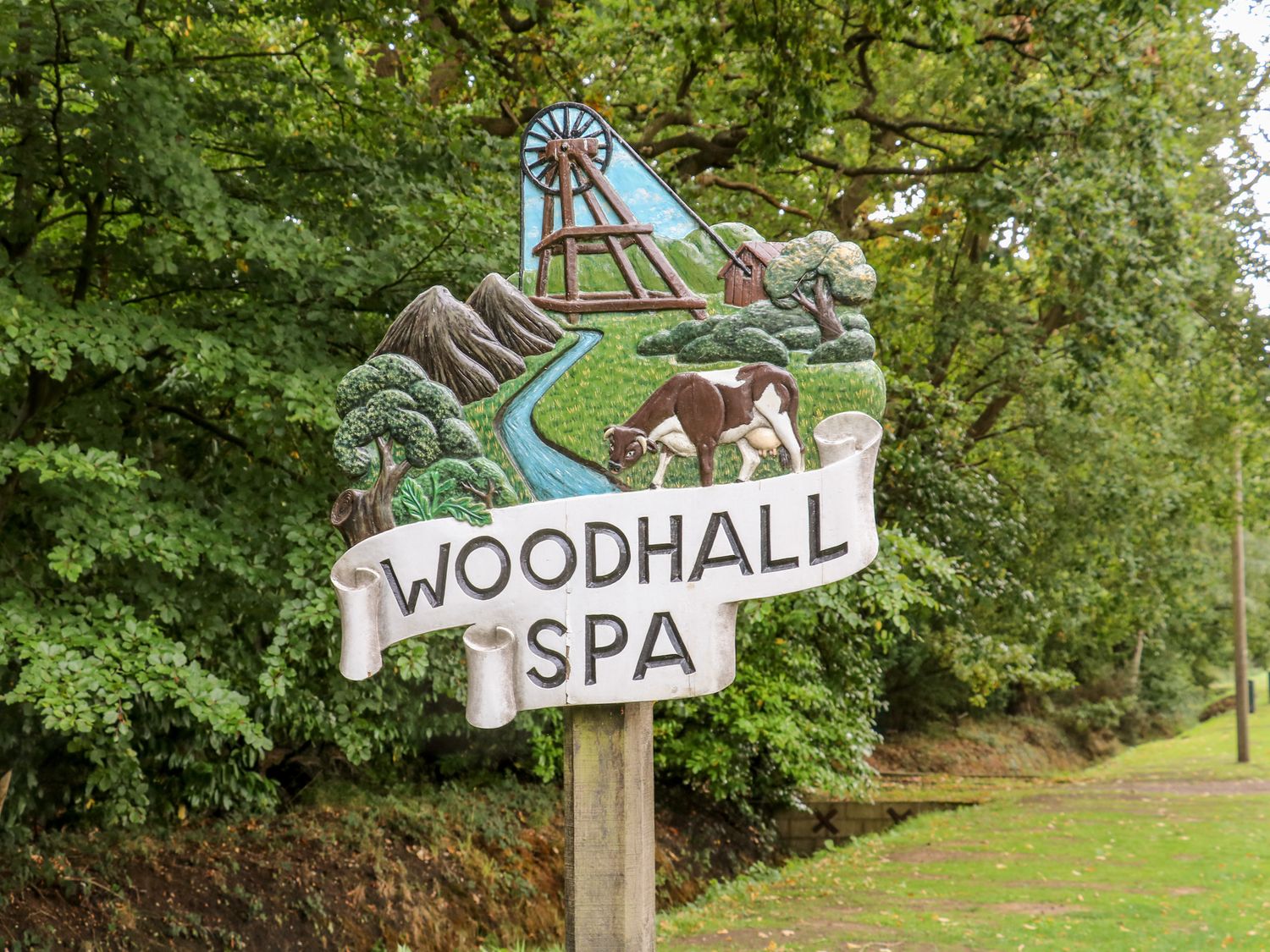 A wooden sign with a cow a river trees mountains and a structure with the text Woodhall Spa in a green outdoor area