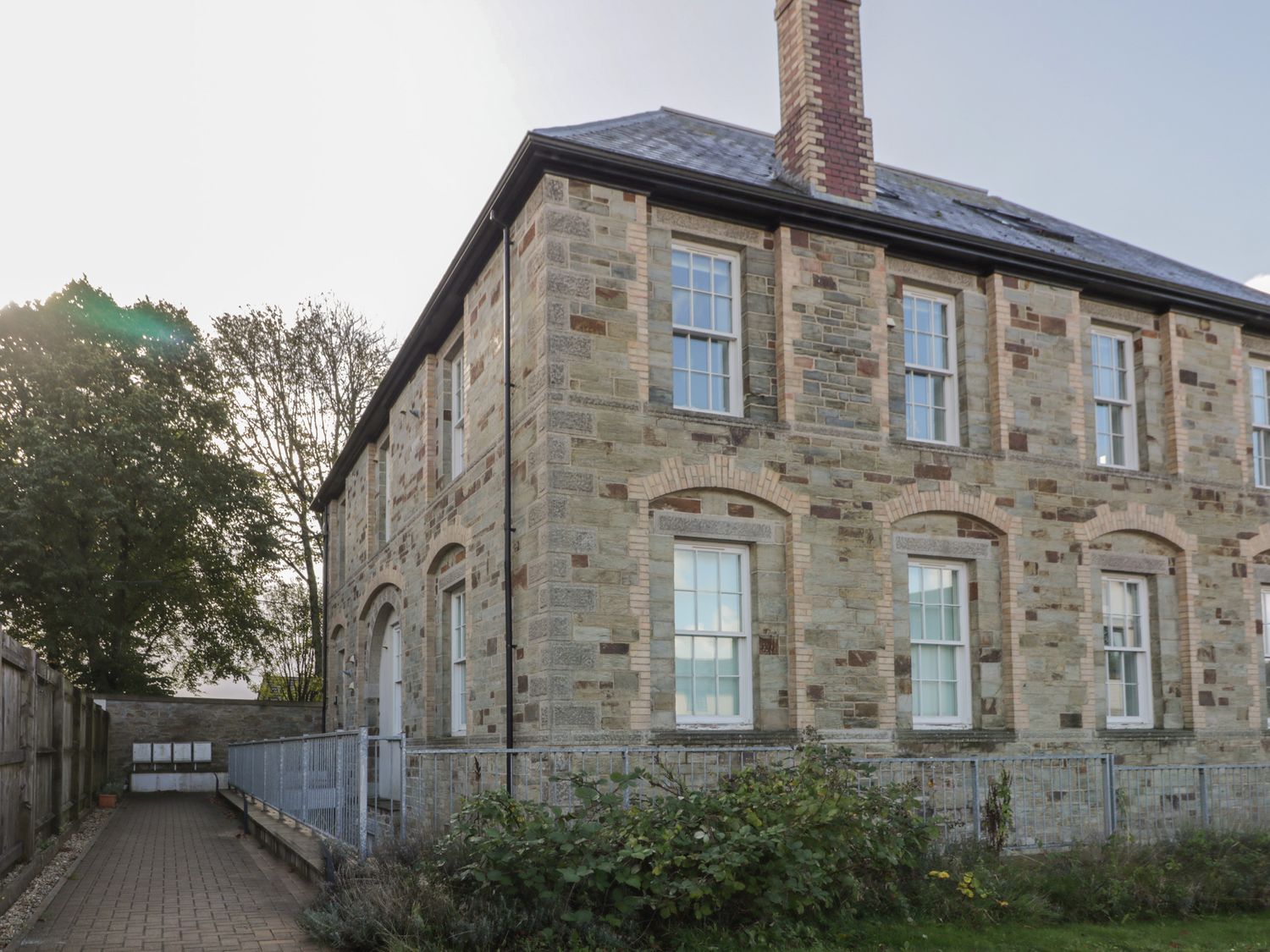 A stone building with multiple white a windows and a chimney with a paved pathway and metal railing at Stores & Supplies in Bodmin