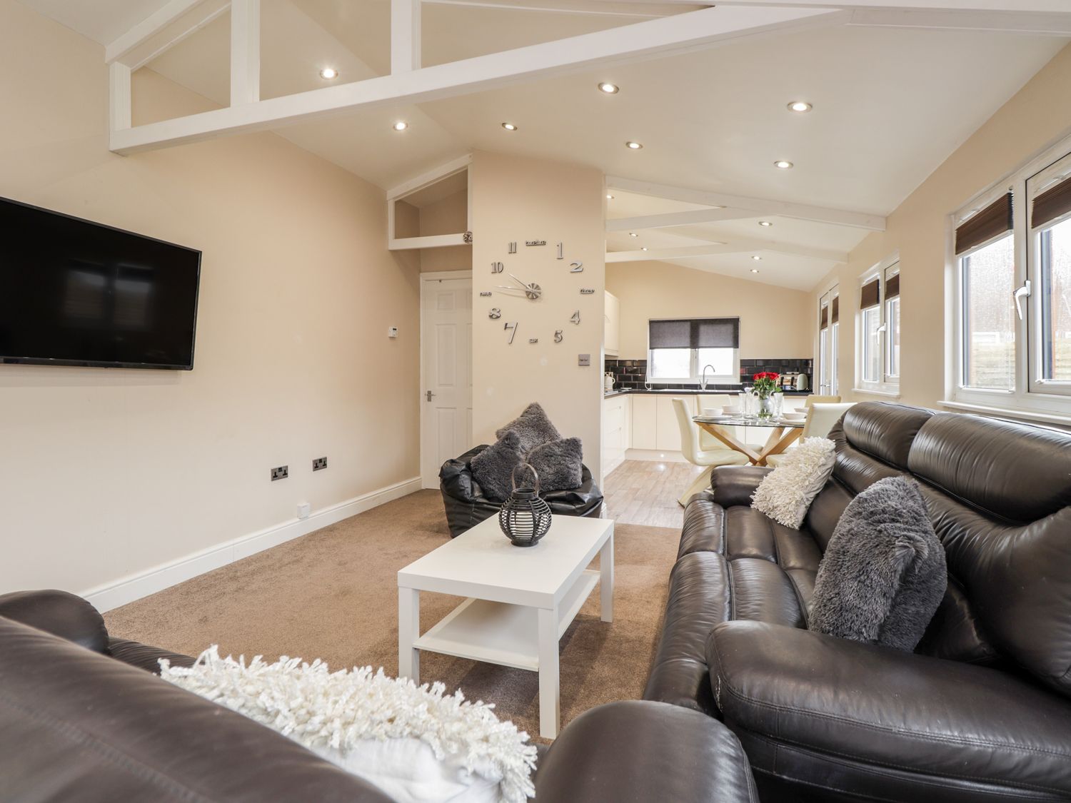 A living room with black leather sofas a white coffee table a wall clock and a wall mounted tv at Sparrows Den Lodge in Winchelsea Beach