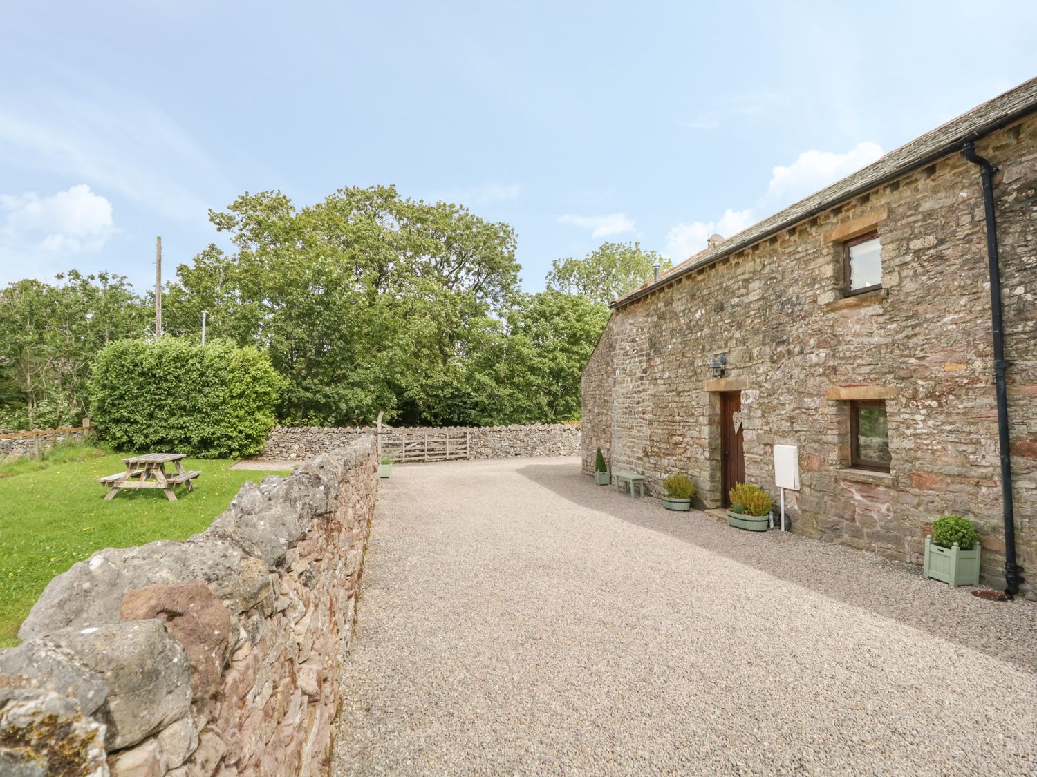 A gravel courtyard beside a stone building with potted plants and a wooden picnic table on grass at Byre Cottage in Great Asby near Appleby-In-Westmorland