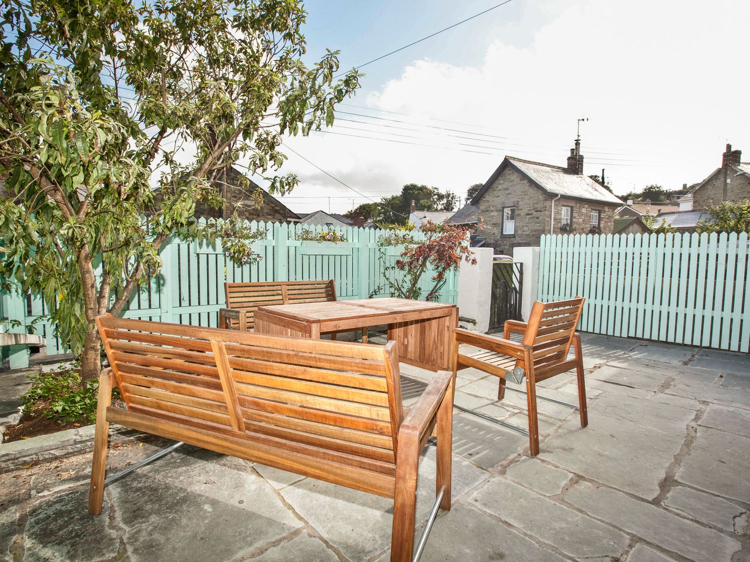 An outdoor patio with wooden benches and chairs around a wooden table near a tree and a pale blue picket fence at 21 British Road in St Agnes