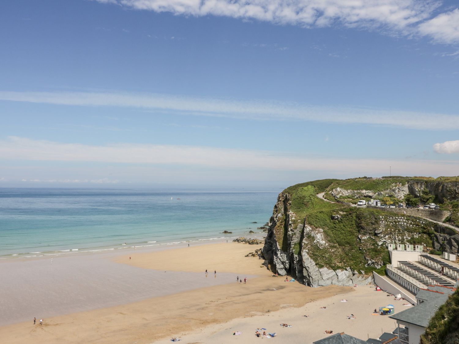 A beach with sand and water next to cliffs with cars parked on top at Two Bare Feet in Newquay