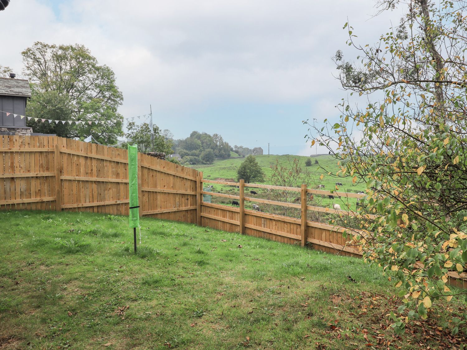 A fenced backyard with grass and a covered green umbrella with a tree to the right and fields in the background at Dove Tail Barn in Kendal