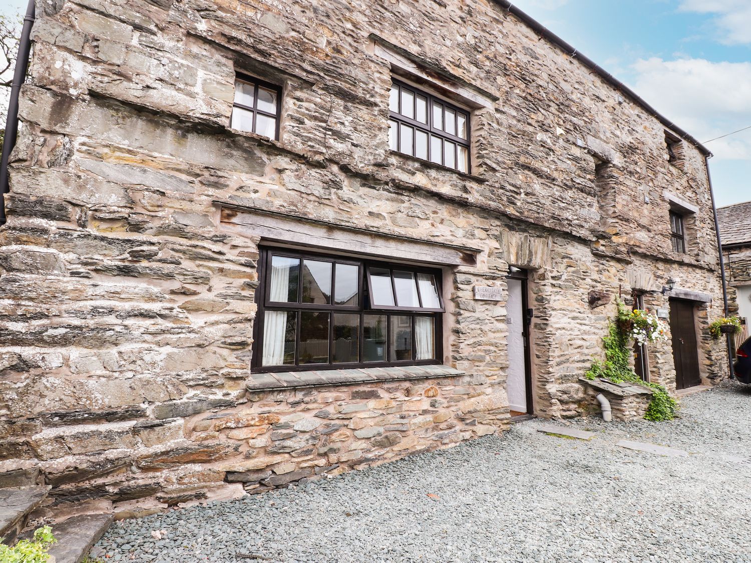 Stone exterior wall of a cottage with windows doors and gravel driveway at Cherry Tree Cottage at Satterthwaite in Hawkshead