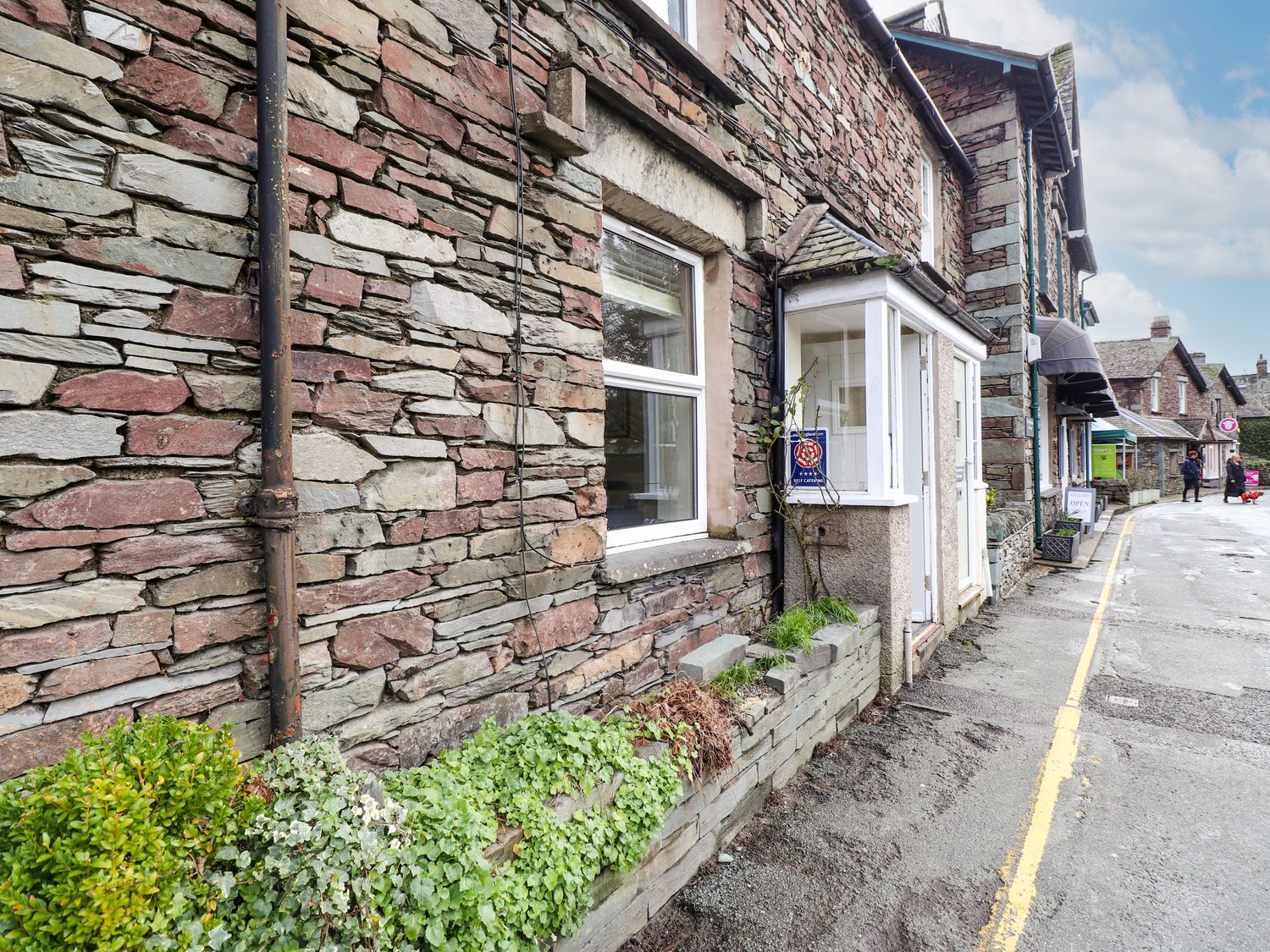 A stone building with a window and a small garden bed along a street with people in the distance at Becksteps Cottage in Grasmere