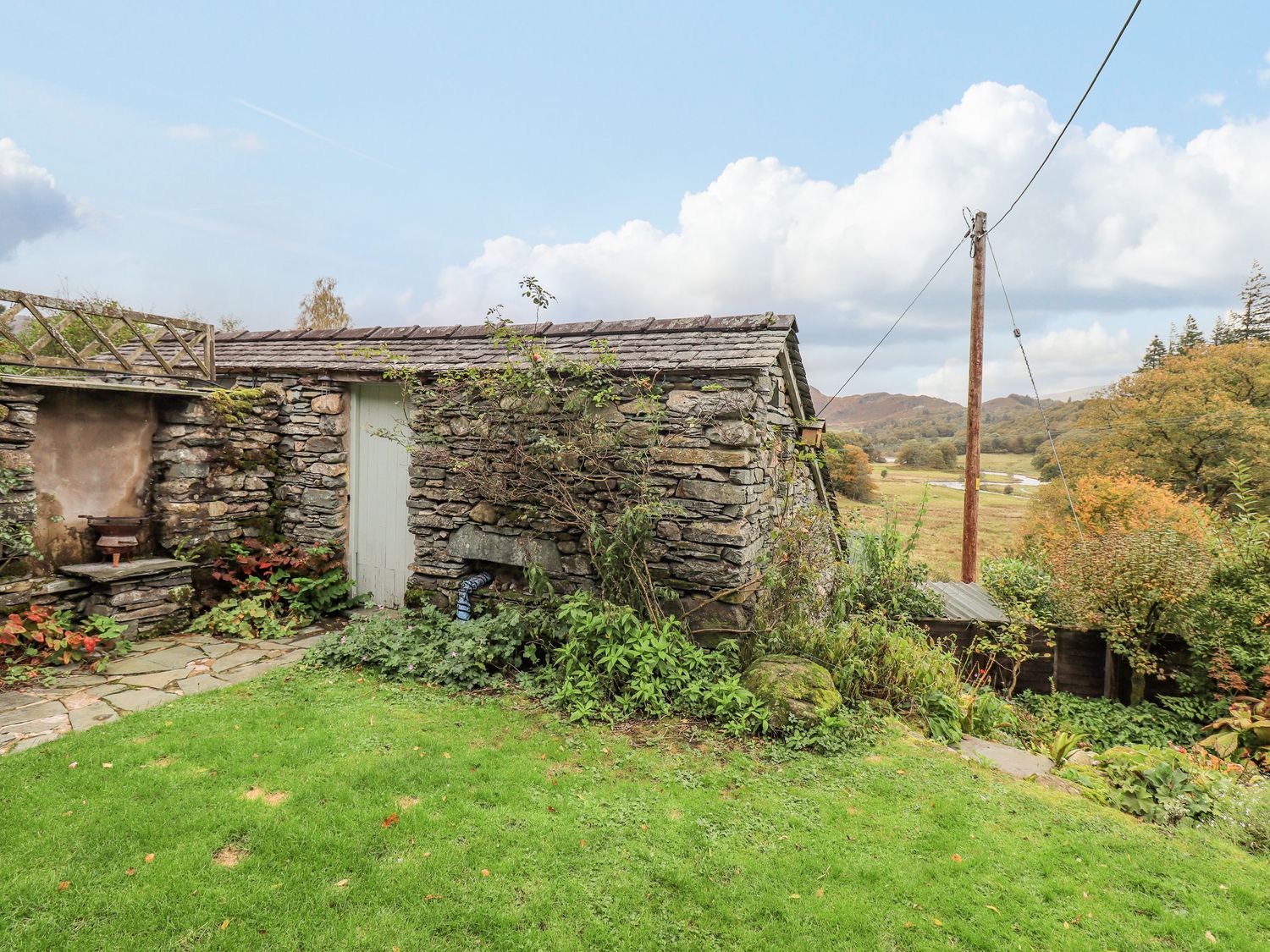 A stone garden shed with plants around and a green lawn with hills and trees in the background at Park Cottage in Ambleside