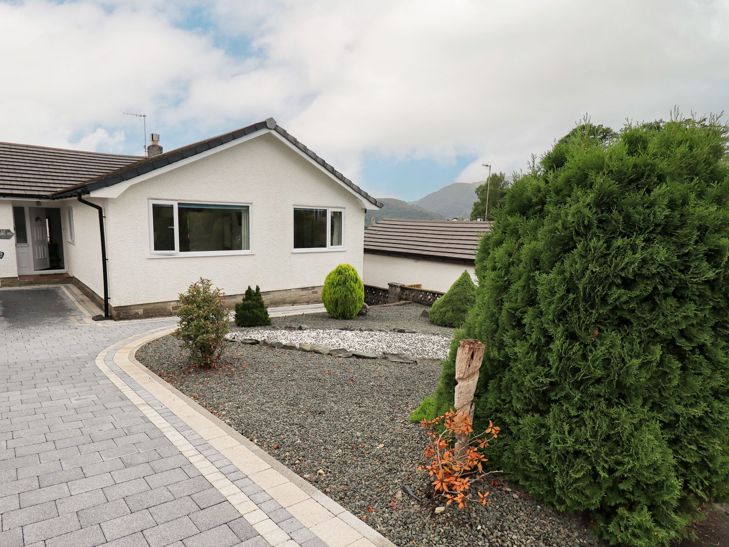 A house with a paved driveway and landscaped garden with various shrubs and mountains in the background at Swirl How in Ambleside