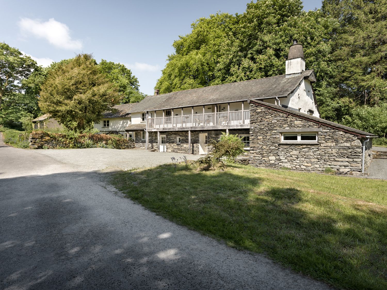 A stone and white two-story house with wooden balcony surrounded by trees and grass at High Lindeth in Bowness