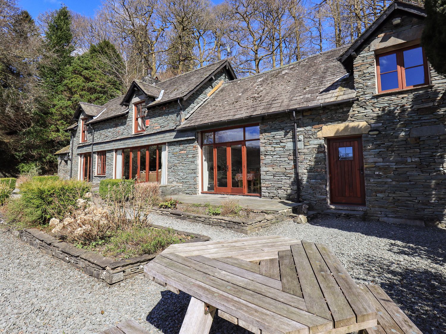 Outside view of a stone house with wooden windows and doors surrounded by trees and garden beds with a wooden picnic table in the foreground at The Stables in Ambleside