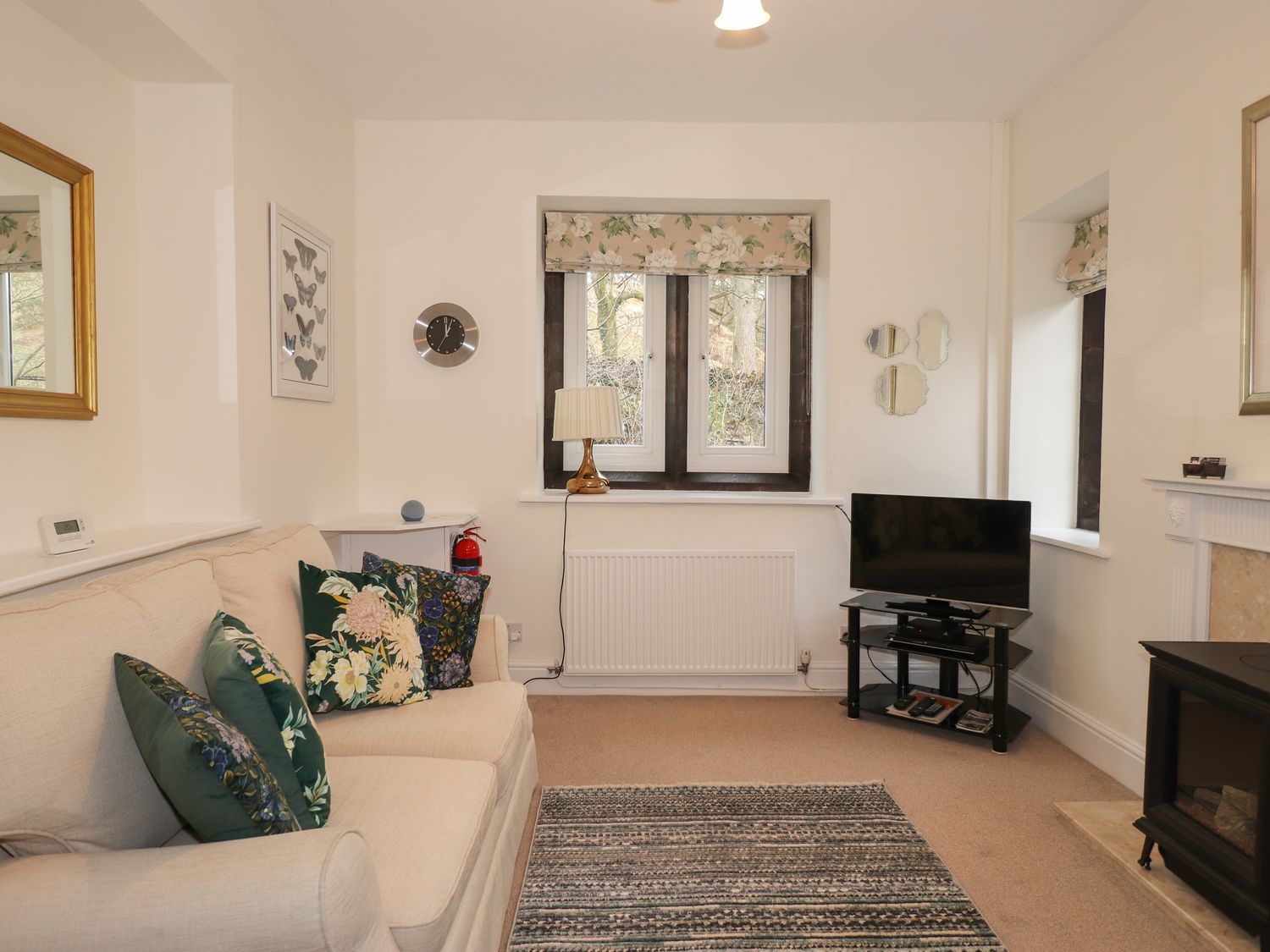 A living room with a beige sofa and floral cushions a TV on a glass stand a window with floral curtains and a fireplace at Larch Cottage in Hawkshead