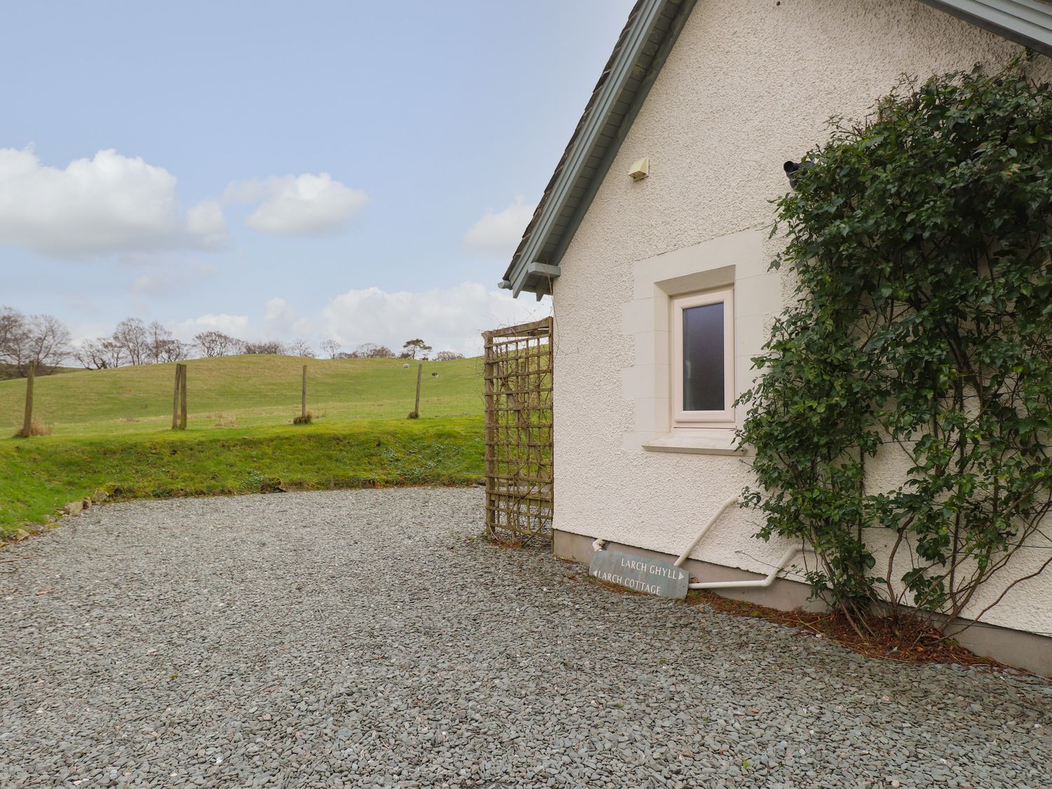 The exterior side of a house with a small window climbing plants a trellis and a gravel area with a grassy hill in the background at Larch Cottage in Hawkshead