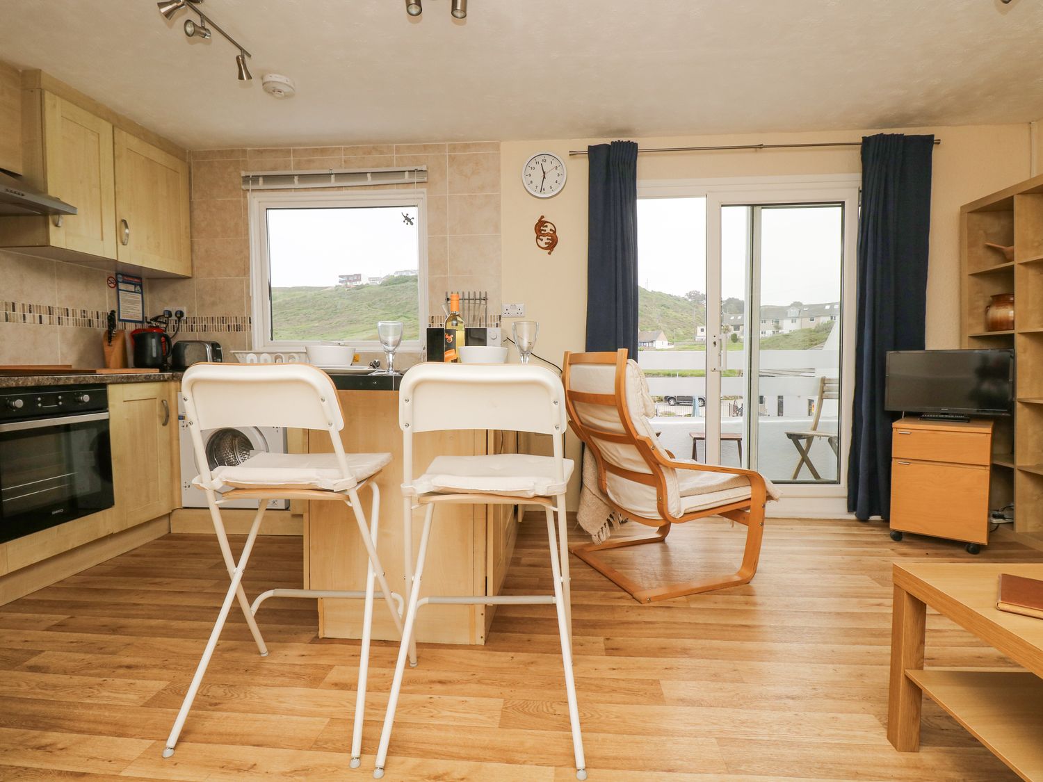 A kitchen and living area with wooden floor white bar stools and a wooden armchair next to sliding glass doors at Gecko Beach in Porthtowan