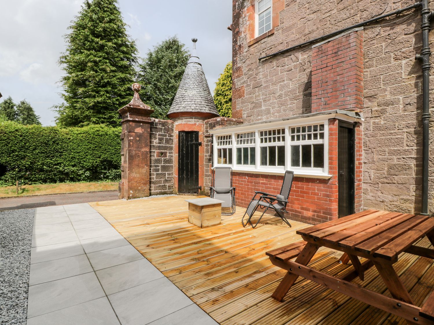 An outdoor patio area with wooden deck chairs a wooden picnic table and a stone building at Apartment 2 in Buchlyvie