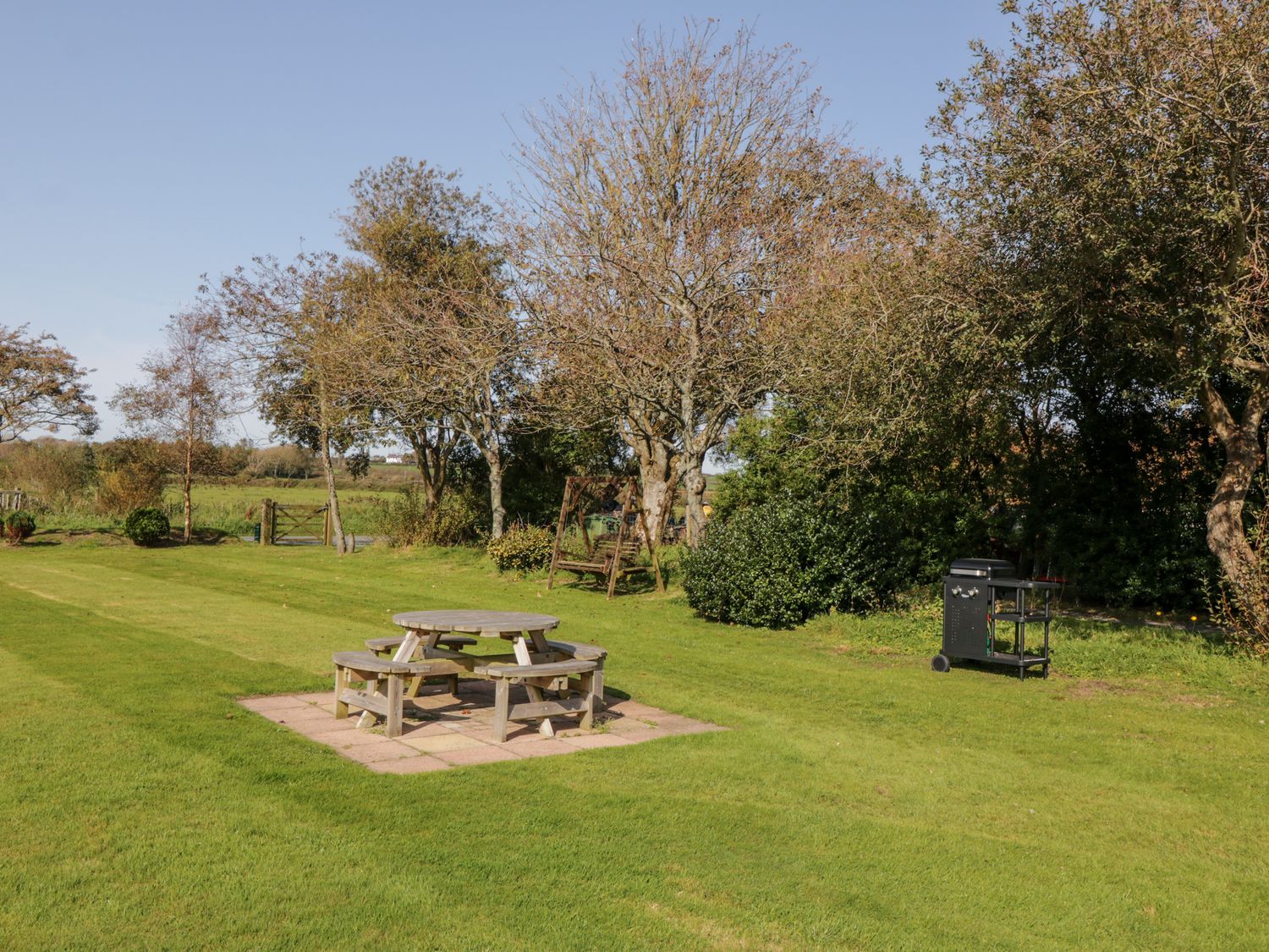 A garden area with a wooden picnic table and benches near trees and a barbecue grill at Penllyn in Newborough