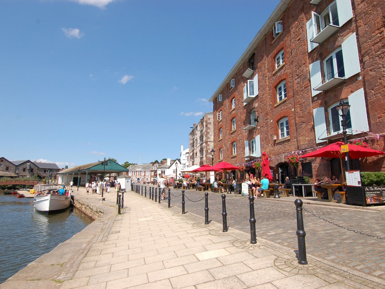 A waterfront promenade with people sitting at outdoor tables with red umbrellas next to a brick building and a docked boat at Broadclyst Cottage in Ottery St Mary