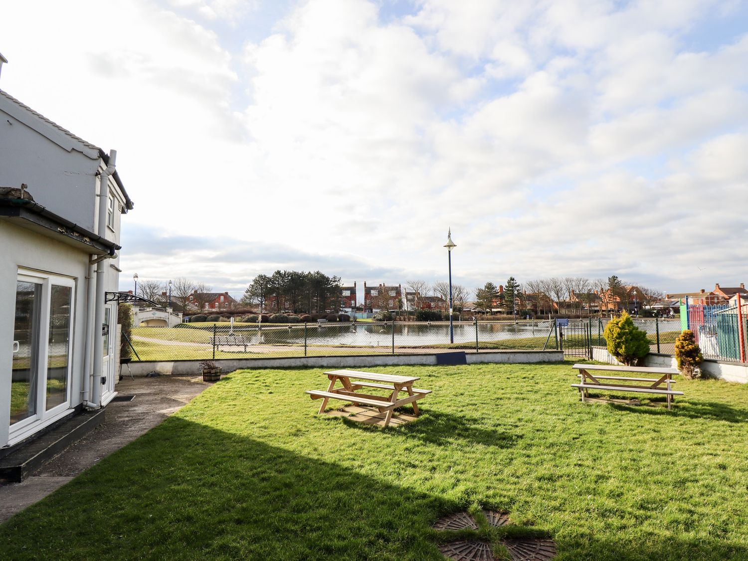 A backyard with green grass and two wooden picnic tables near a fenced pond at Bank House in Mablethorpe