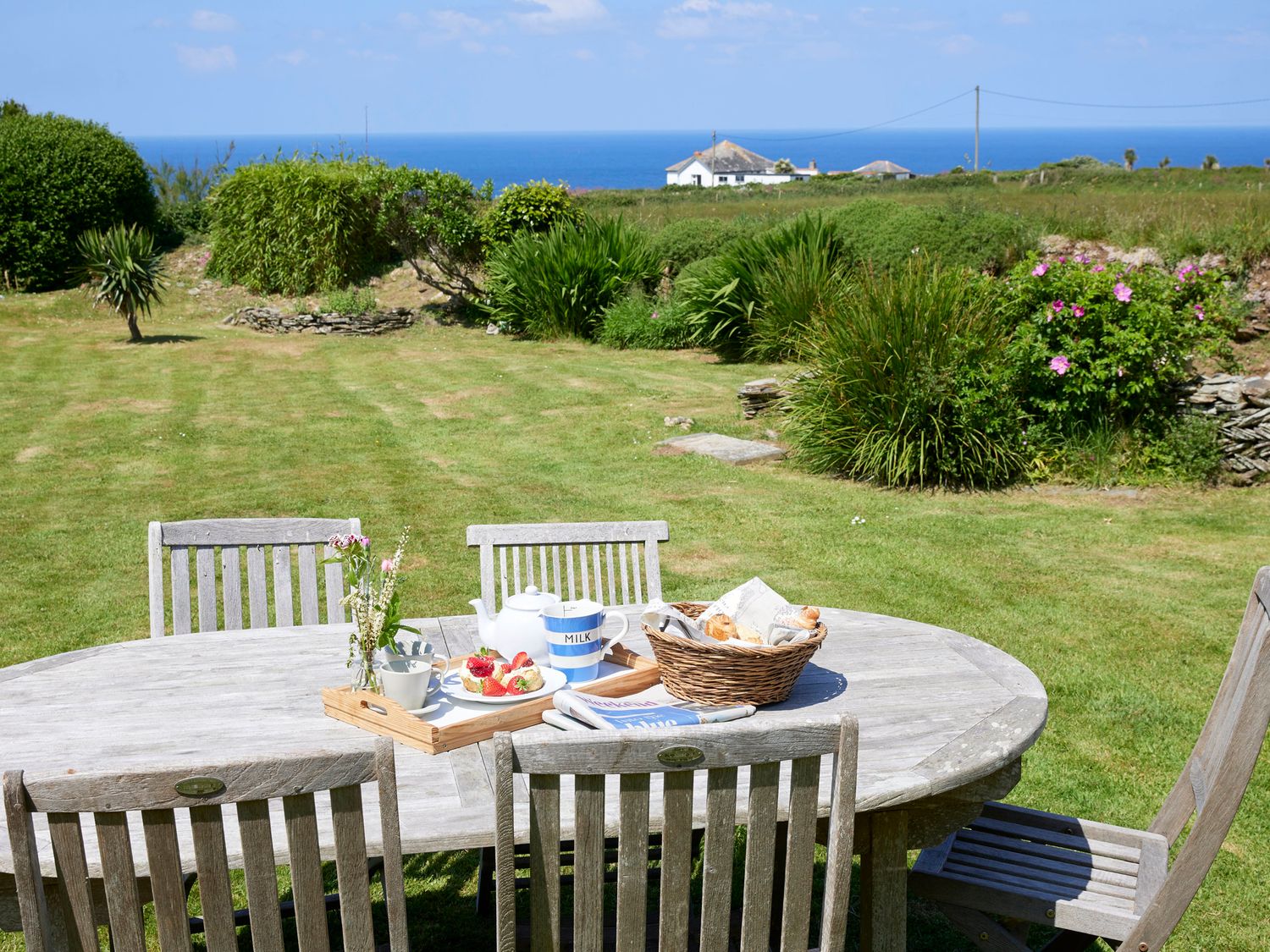 An outdoor wooden table with chairs on a lawn overlooking shrubbery and the ocean at Merlins Cottage in Tintagel