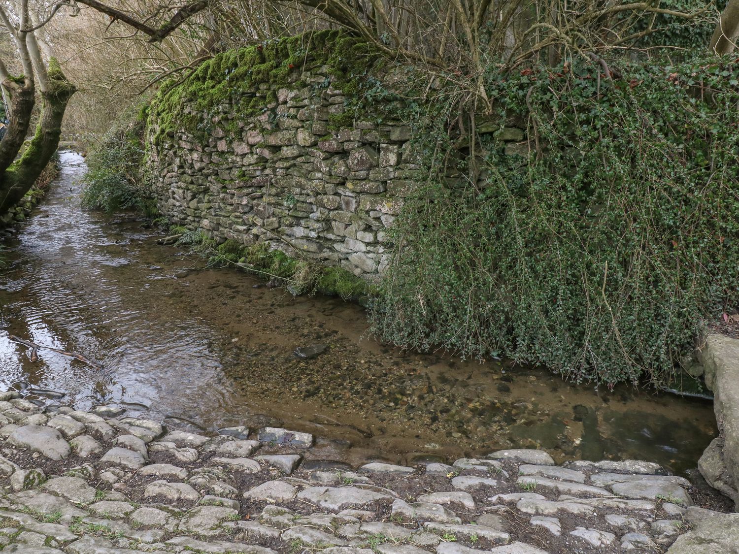 A stone wall with moss beside a shallow stream bordered by a cobblestone path at Brookhouse in Castleton Peak District