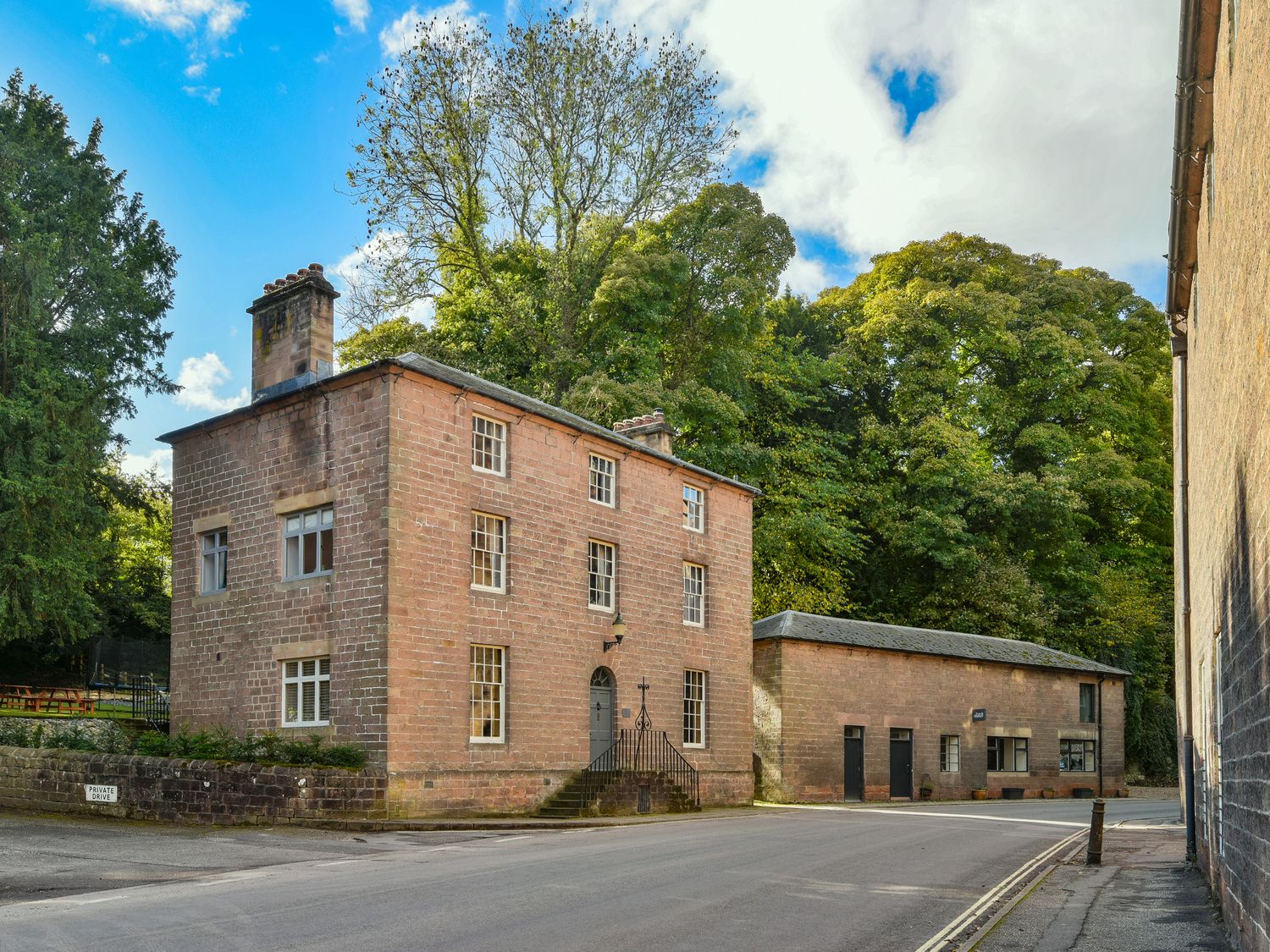 A stone building with multiple windows and a chimney on a road next to another stone building and trees at The Mill Managers House in Cromford