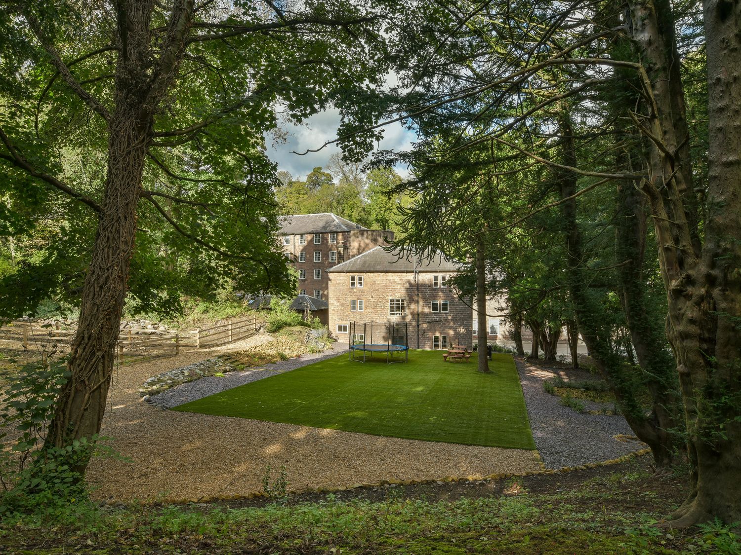 An outdoor area with grass and a trampoline surrounded by trees and stone buildings at The Mill Managers House in Cromford