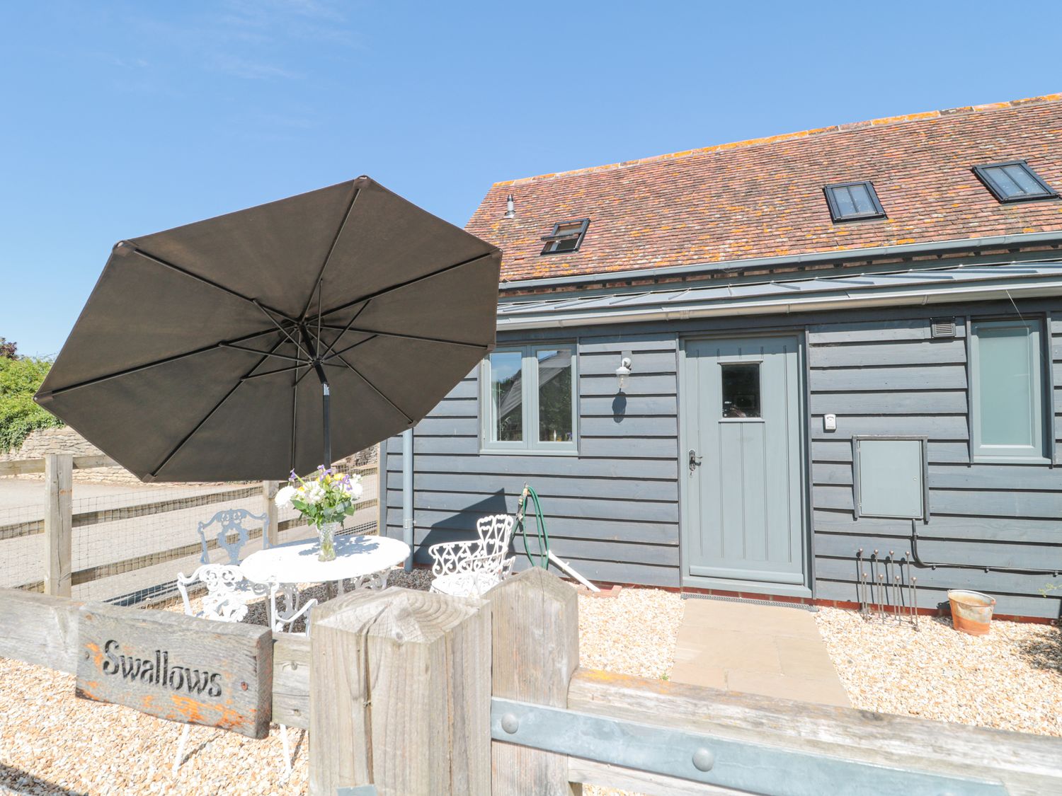 An outdoor patio area with a table, chairs, umbrella, and flowers in front of a gray wooden building at Swallows, 1 The Old Stables in Stanton Harcourt near Witney