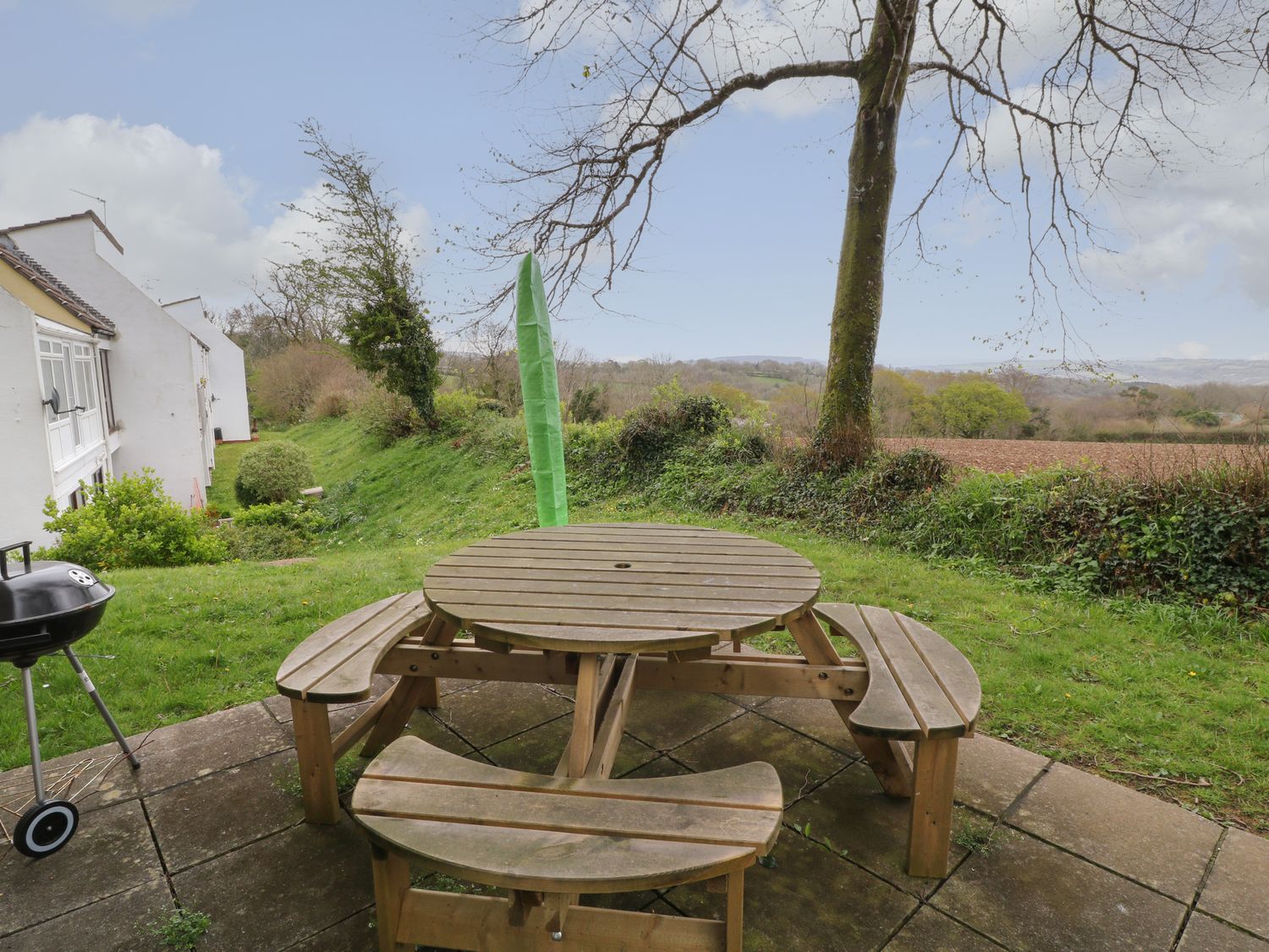 A wooden round picnic table with benches on a patio near a tree and a grassy area at Hembry Meadows in Honicombe Park near Gunnislake