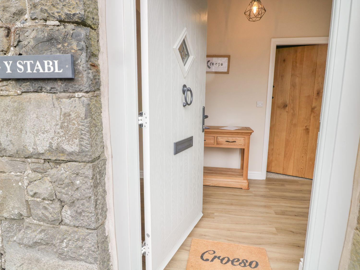 An open white door revealing a wooden floor inside a hallway with a wooden console table a hanging light and a door mat with Croeso at Y Stabl in Llanddoged near Llanrwst
