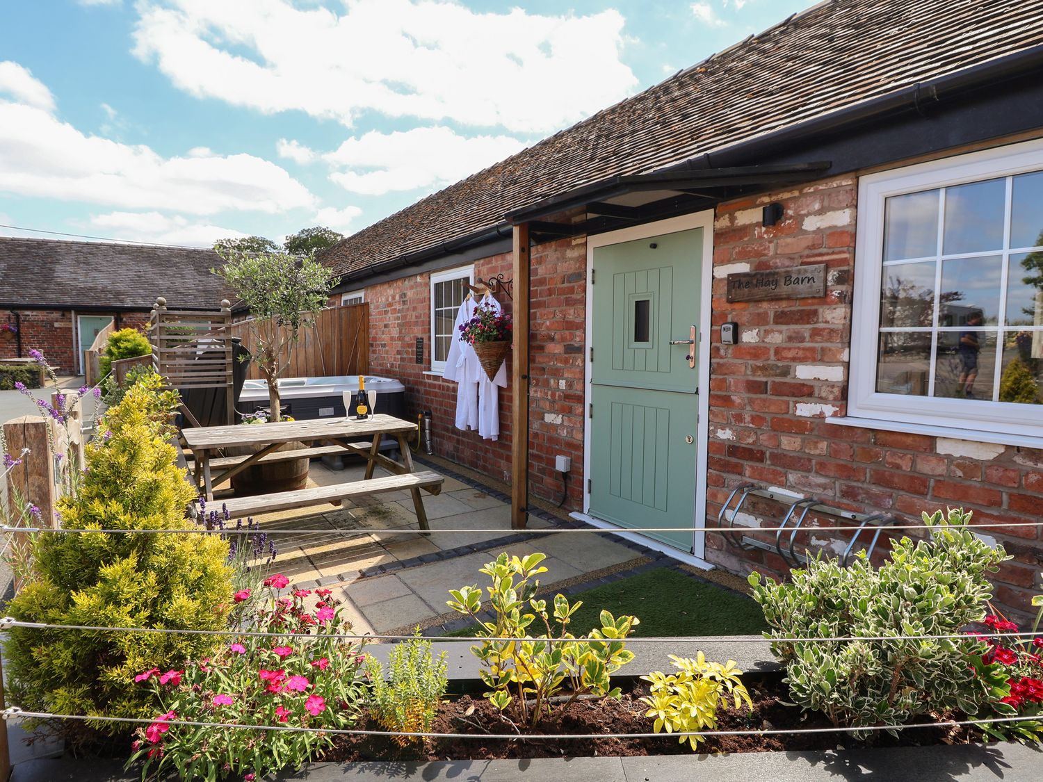 An outdoor patio area with a wooden picnic table a hot tub flowers and a green door on a brick building at The Hay Barn in Chester