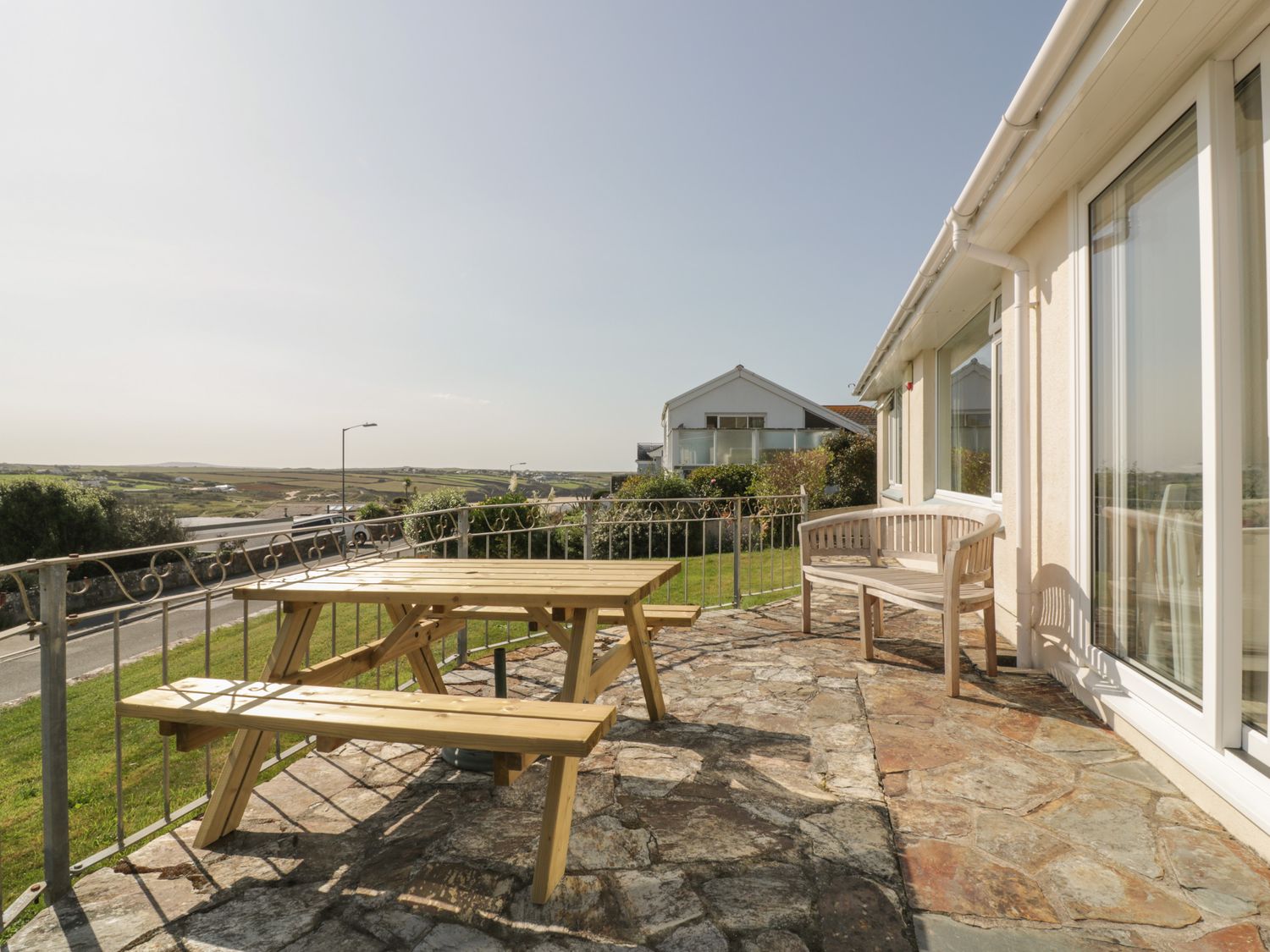 An outdoor stone patio with a wooden picnic table and bench along a cream house with glass sliding doors at Gwel An Mor in Newquay