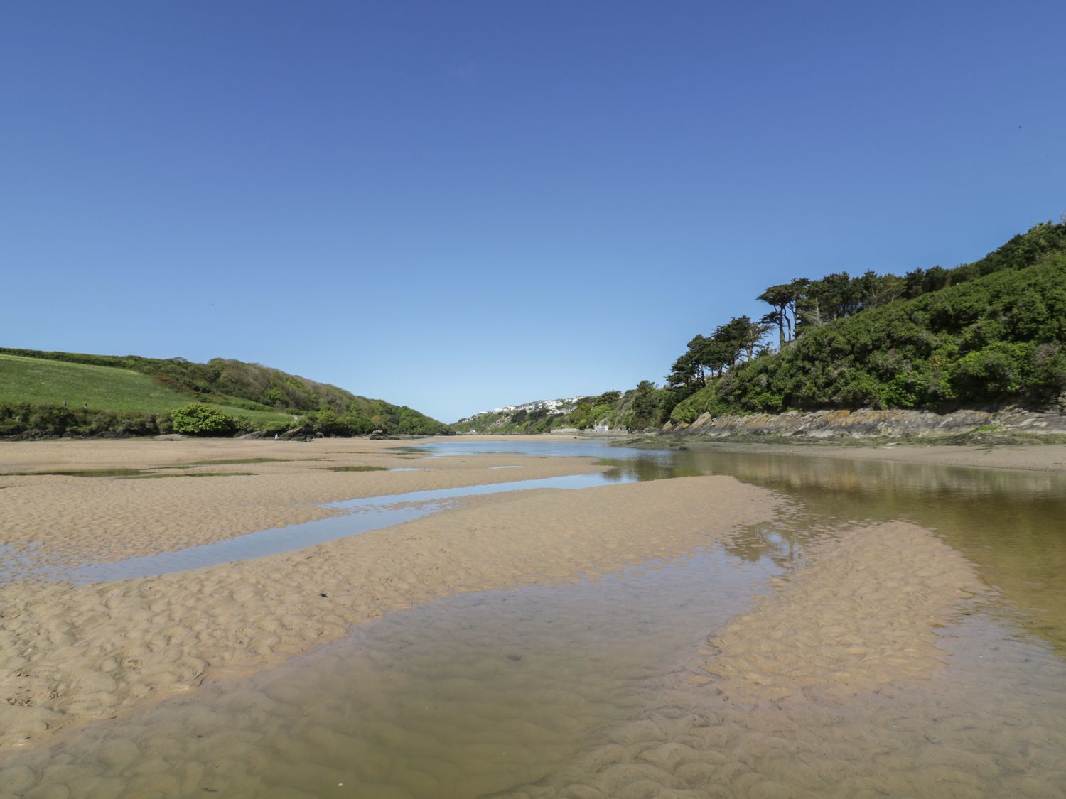 A sandy riverbank with water pools surrounded by green hills and trees under a clear blue sky at Gwel An Mor in Newquay