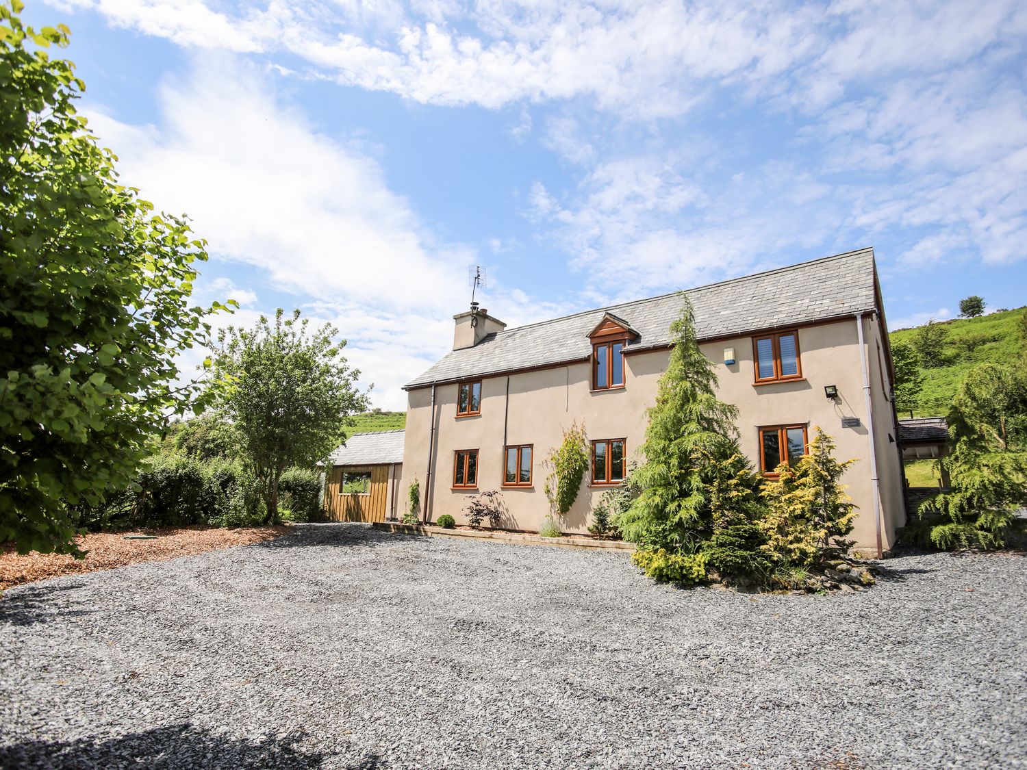 A gravel driveway with trees and shrubs around a two-story house under a blue sky at Hafotty Gelynen Maerdy near Corwen
