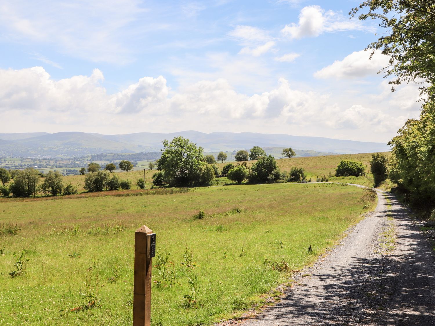 A gravel road running alongside a field with trees and hills in the background at Hafotty Gelynen in Maerdy near Corwen
