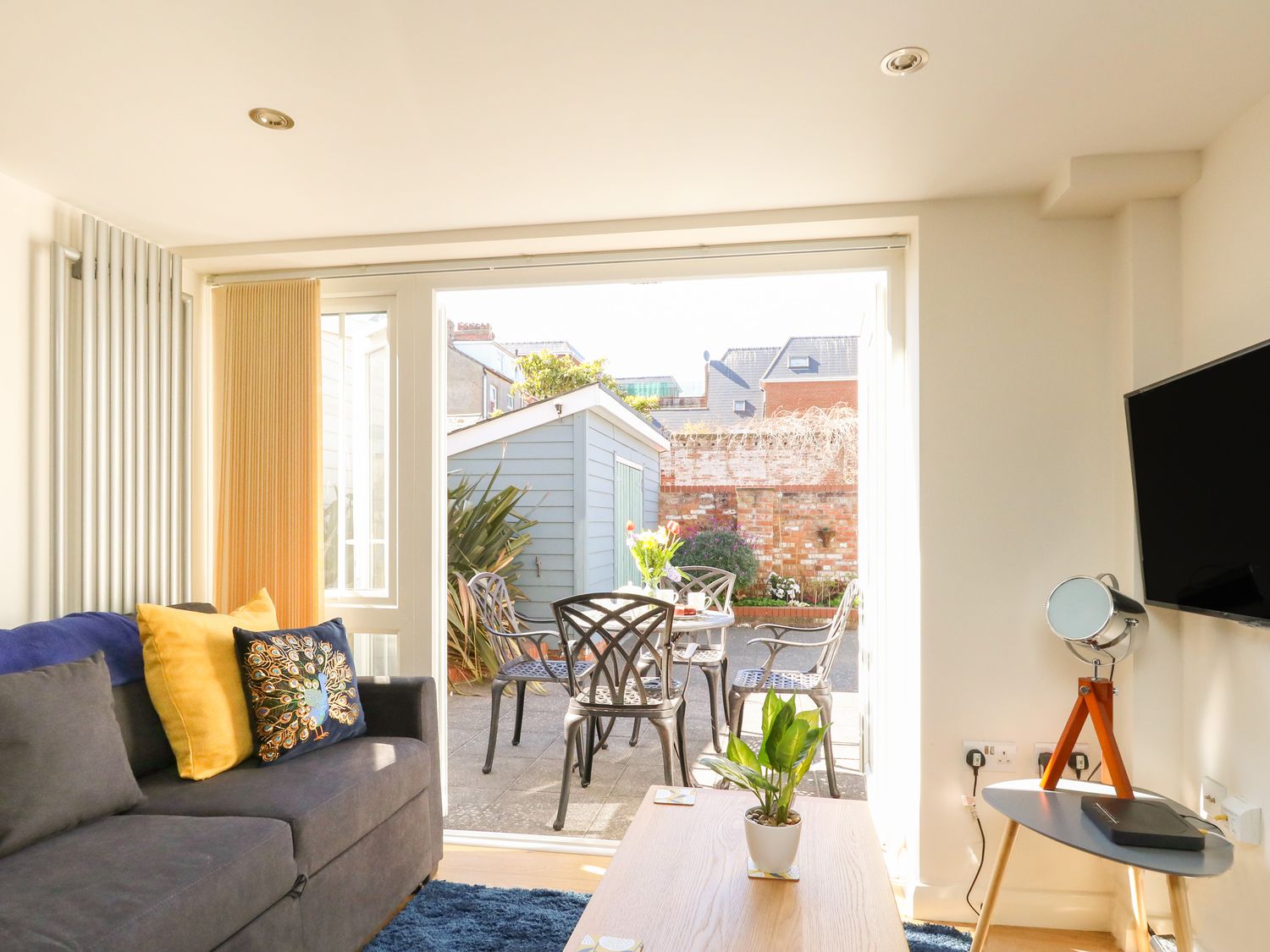 A living room with a gray sofa yellow and patterned cushions a small table with a plant and a television on the wall at Park Mews in Weymouth
