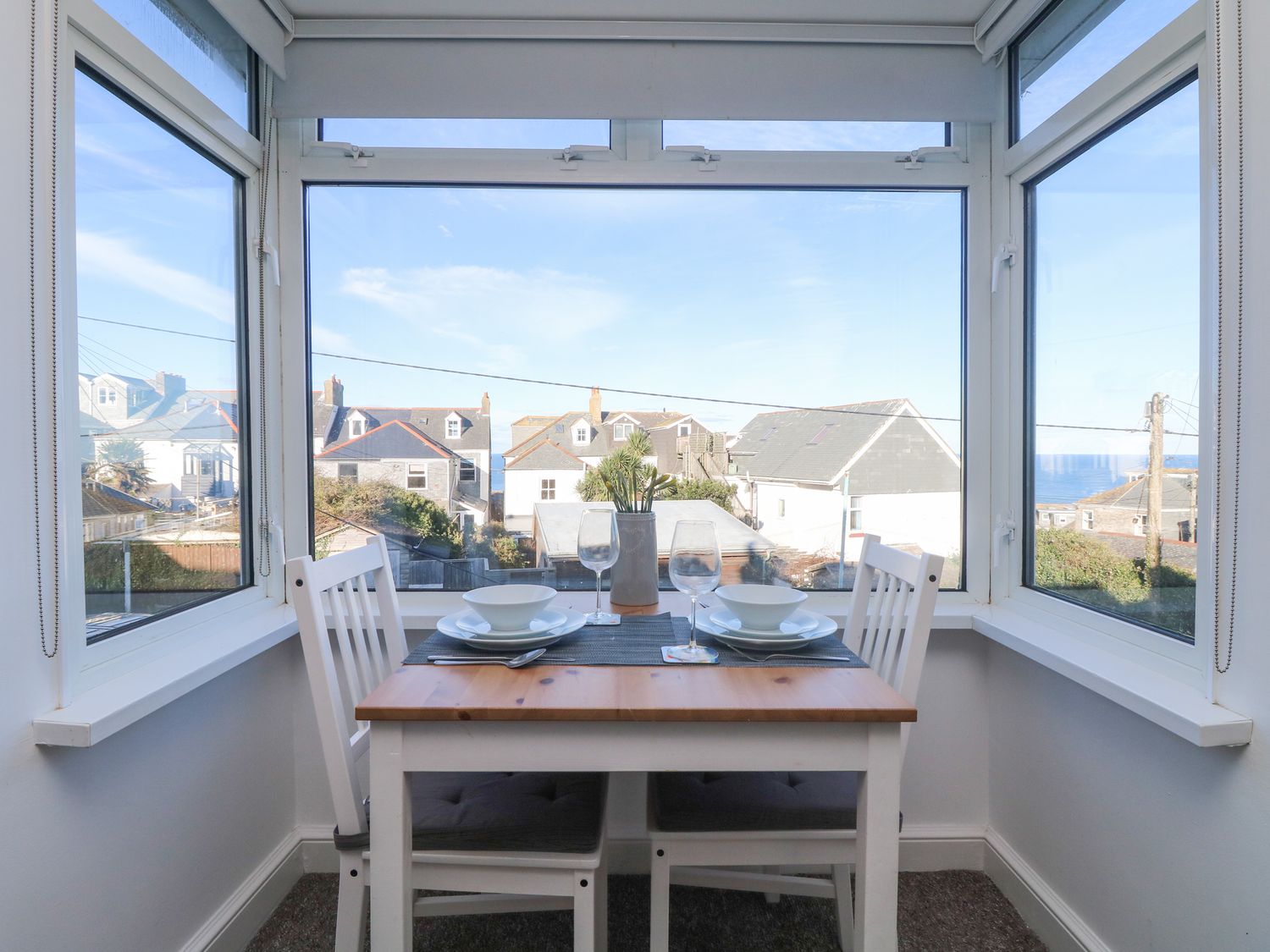 A dining table set for two by a bay window with views of houses and the sea at The Beach Hut in St Ives